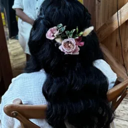 Upięcie z kwiatami na ciemnych, długich włosach, widok z tyłu. Kobieta siedzi na drewnianym krześle. W tle fragment wnętrza z drewnianymi elementami.