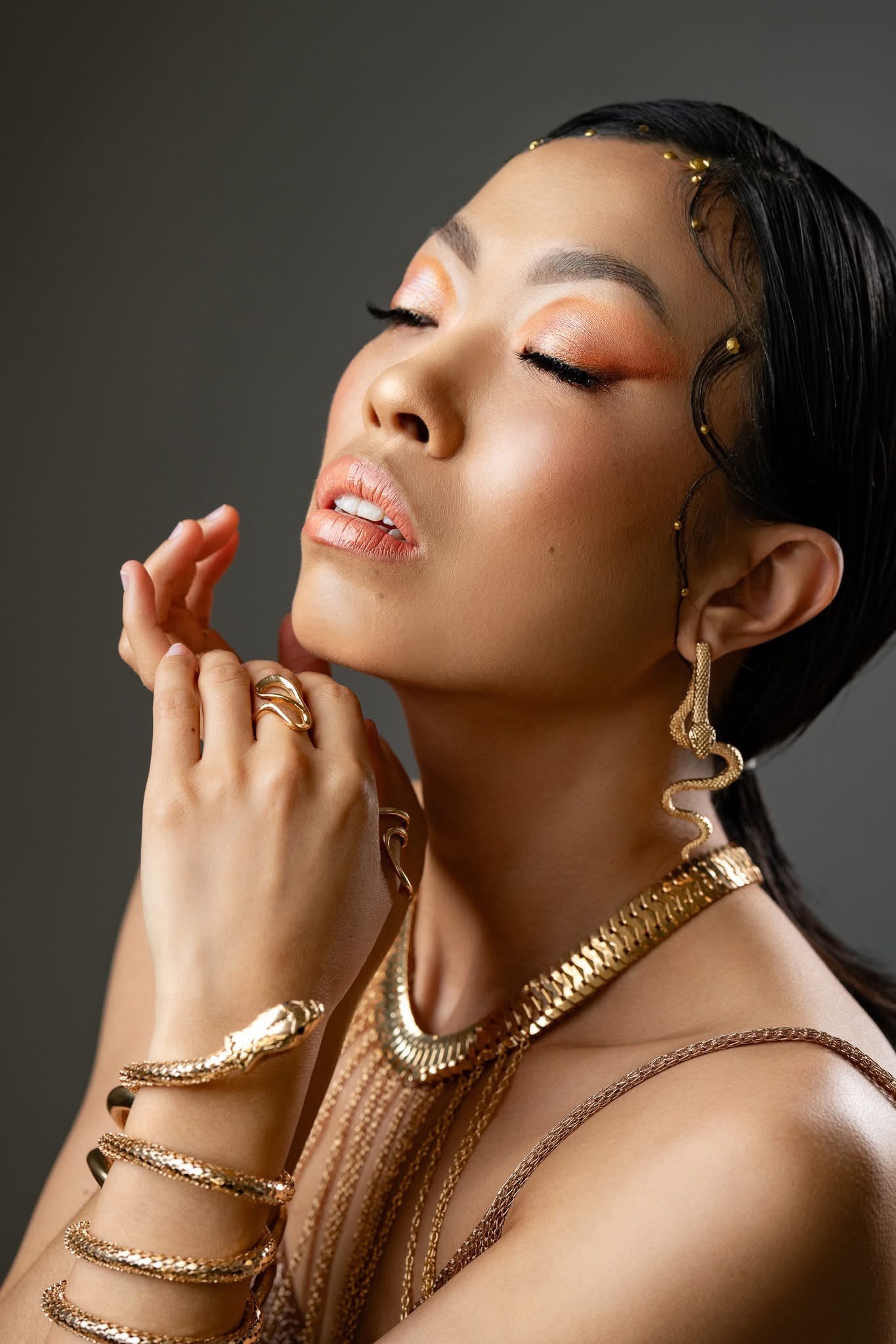 Portret kobiety z zamkniętymi oczami, w bogatej złotej biżuterii w stylu węża, z intensywnym pomarańczowo-złotym cieniowaniem powiek i błyszczącymi ustami, na gładkim szarym tle.