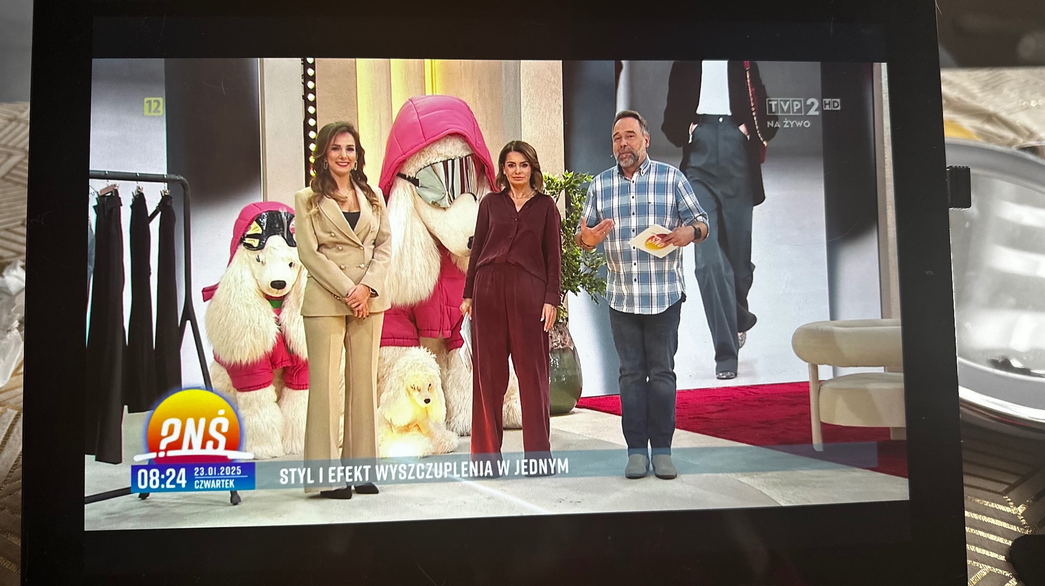 Prezenterki telewizyjne i prezenter w studio telewizyjnym podczas programu o modzie i stylu, obok manekiny psów w ubraniach i wieszak z ubraniami