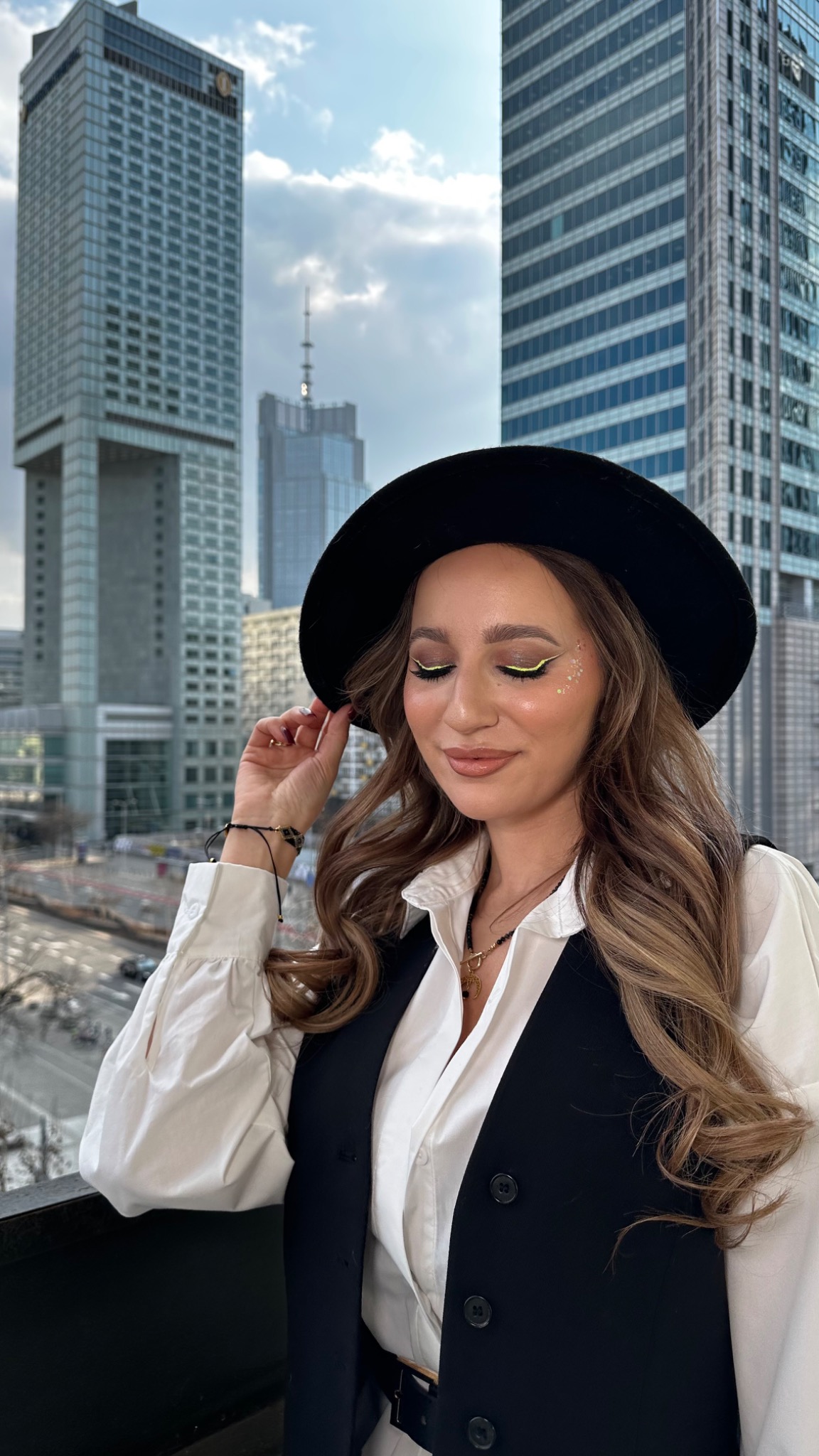 Portret kobiety w czarnym kapeluszu z delikatnym, neonowym makijażem oczu i błyszczącymi drobinkami na policzku, na tle panoramy miasta.