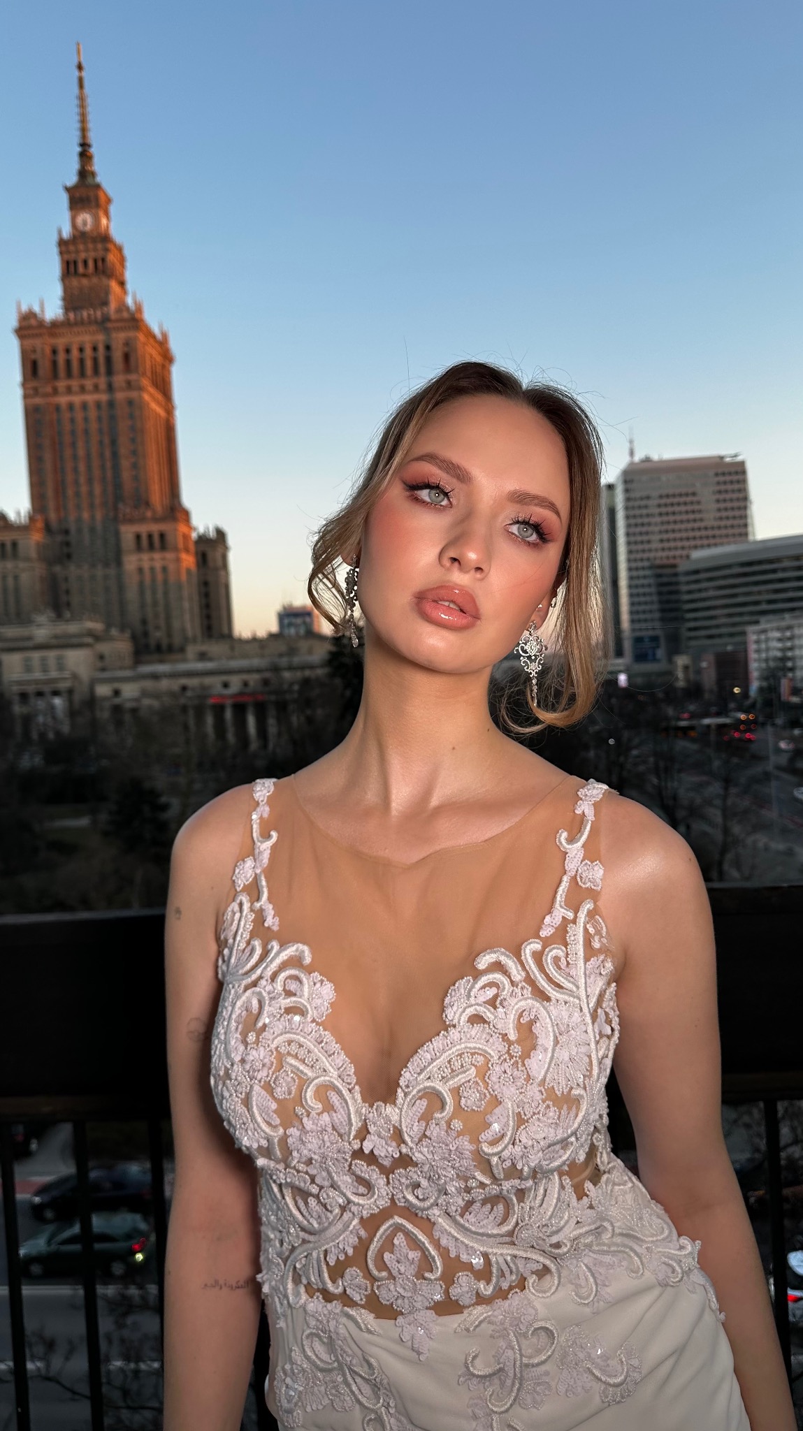 Portret kobiety z delikatnym, różowym makijażem oczu i ust, w ozdobnej sukni na tle panoramy Warszawy o zmierzchu.