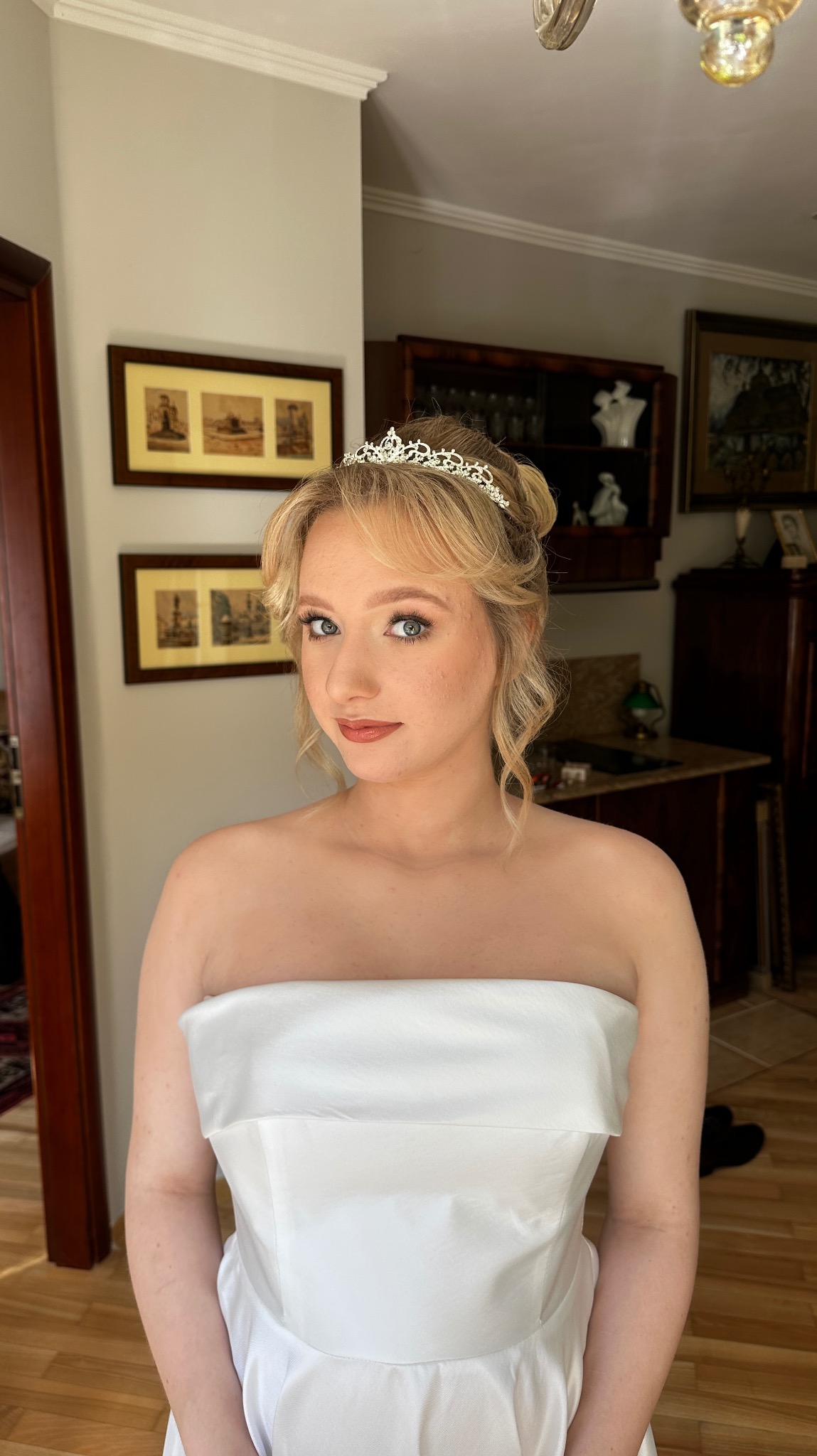 Portret kobiety w białej sukni i tiarze, z delikatnym makijażem i upiętymi blond włosami, na tle wnętrza z obrazami w ramkach.