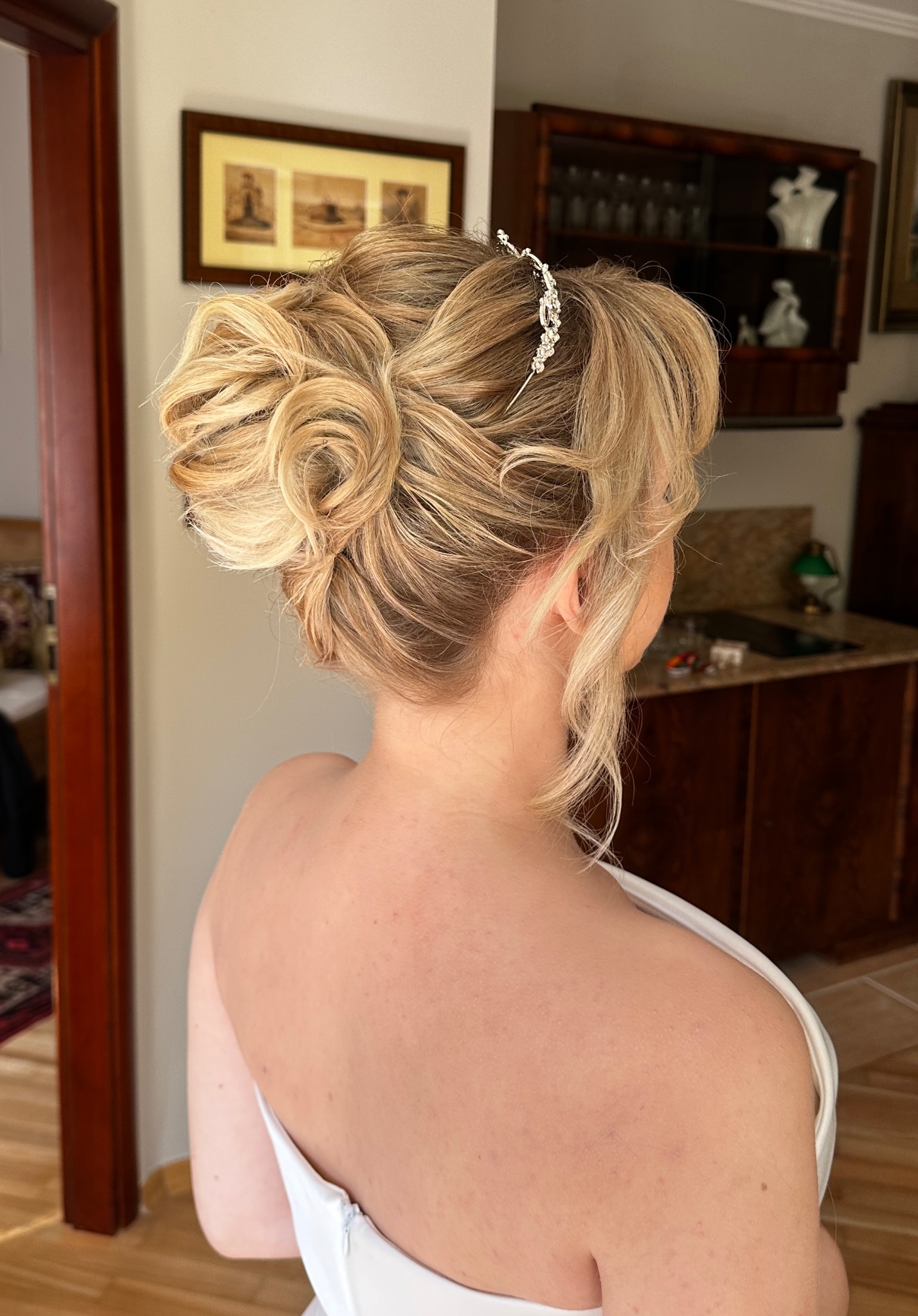 Elegancka fryzura ślubna z upięciem i ozdobną spinką, widok z tyłu na tle wnętrza z drewnianymi meblami.
