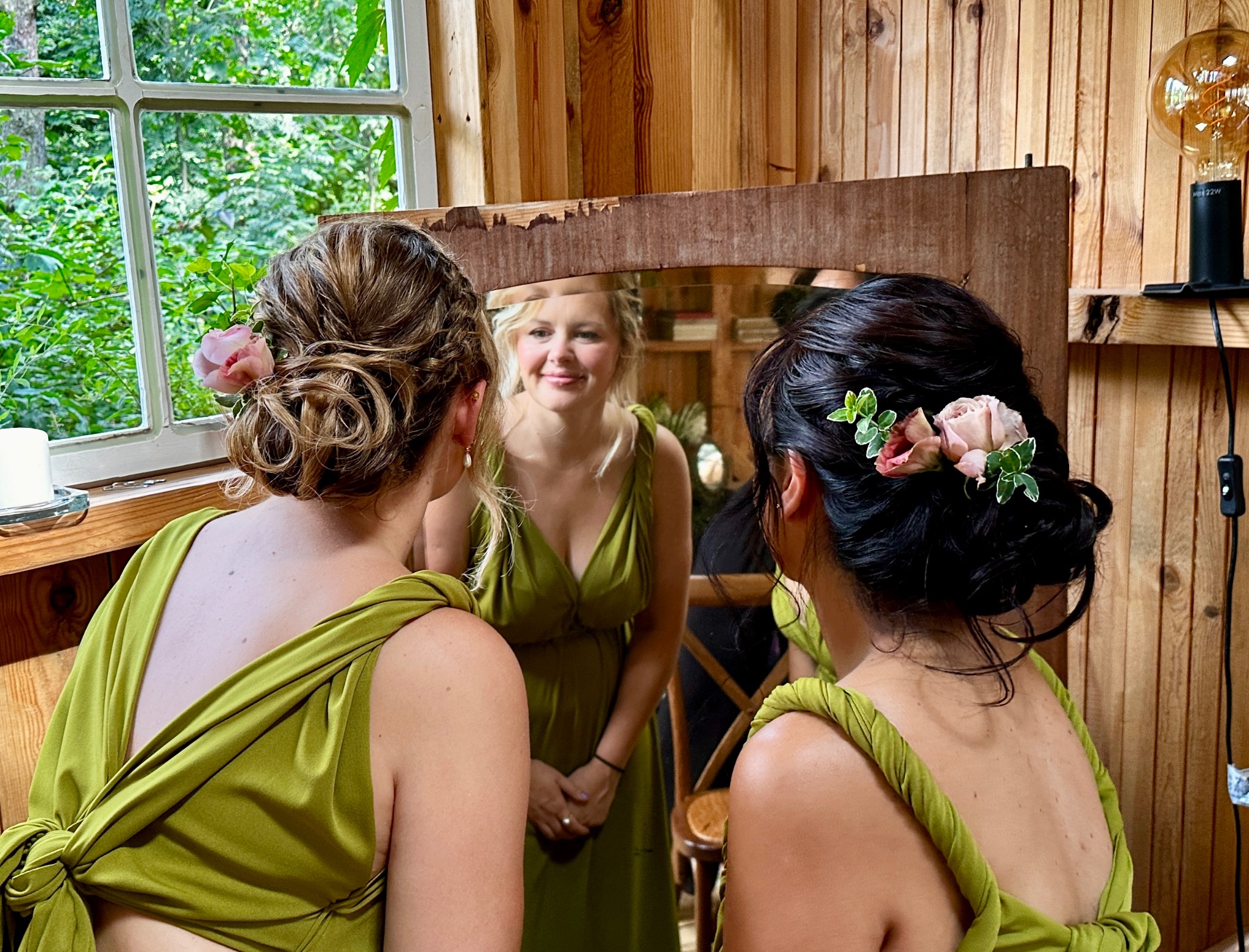 Dwie kobiety z upięciami z kwiatów w odcieniach różu i zieleni, widoczne od tyłu, odbijają się w rustykalnym lustrze w drewnianym wnętrzu.