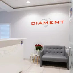 Jasne wnętrze poczekalni z recepcją, szarą sofą, bukietem czerwonych róż i logo 'DIAMENT' na ścianie.