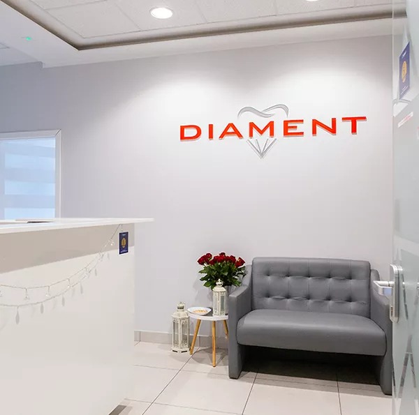 Jasne wnętrze poczekalni z recepcją, szarą sofą, bukietem czerwonych róż i logo 'DIAMENT' na ścianie.