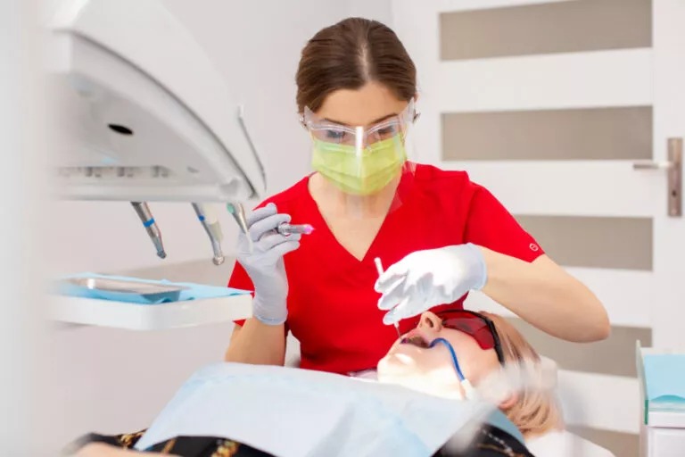 Lekarz w czerwonym uniformie, z maską i okularami ochronnymi, wykonuje zabieg u pacjentki leżącej na fotelu dentystycznym, używając strzykawki z różowym płynem.