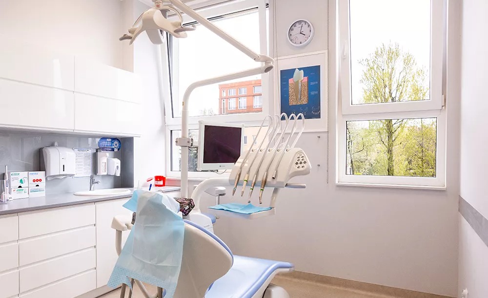 Jasne wnętrze gabinetu z nowoczesnym fotelem dentystycznym, lampą operacyjną, narzędziami i umywalką; na ścianie plakat z przekrojem zęba i zegar.