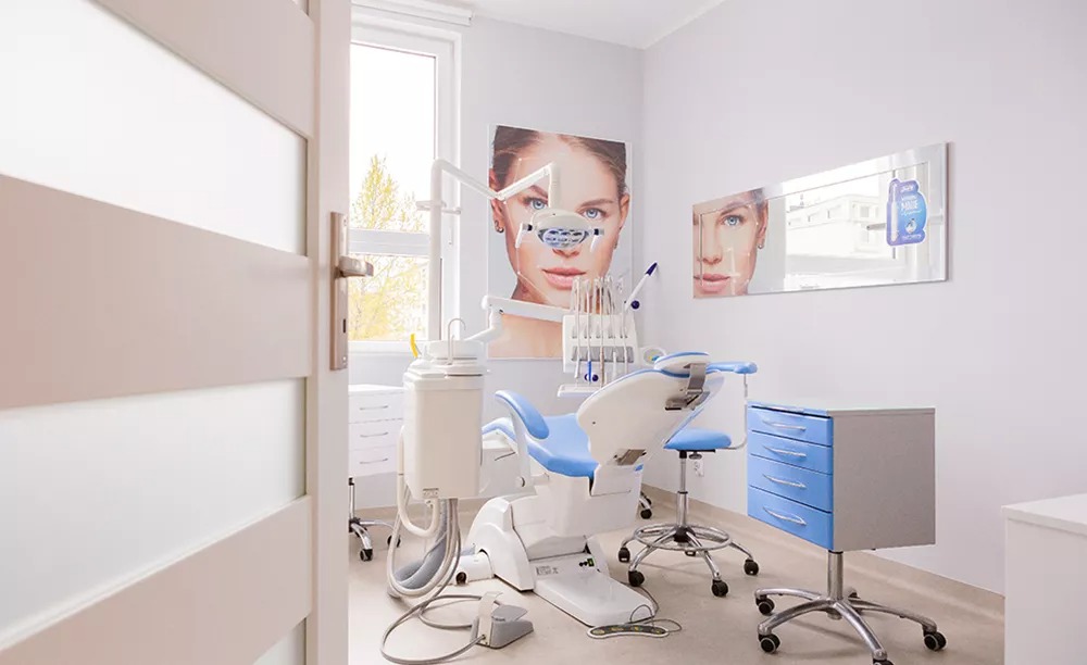 Jasny gabinet z niebieskim fotelem dentystycznym, lampą i szafką na kółkach, widok przez uchylone białe drzwi, na ścianie grafika z twarzą kobiety.