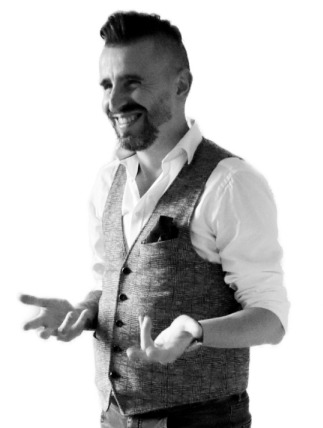 Uśmiechnięty mężczyzna w kamizelce, z charakterystyczną fryzurą, gestykulujący otwartymi dłońmi na jasnym tle.