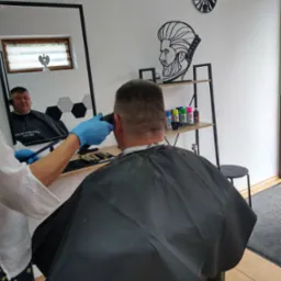 Strzyżenie męskie maszynką, widok z tyłu na klienta w czarnej pelerynie, detale wystroju salonu: zegar z motywem fryzjerskim i półka z kosmetykami do włosów.