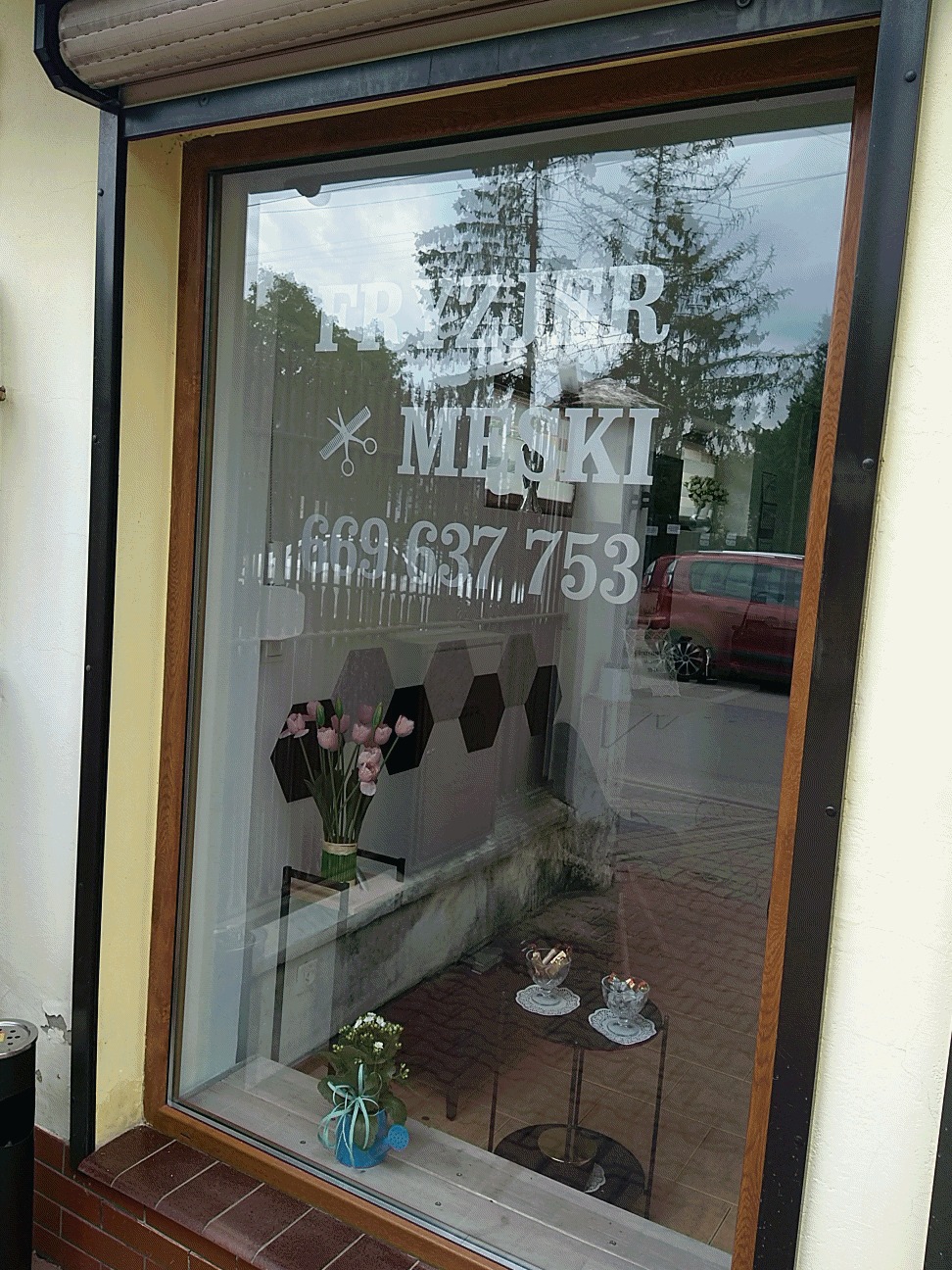 Okno zakładu z napisem 'FRYZJER MĘSKI', numerem telefonu i grafiką nożyczek, dekorowane kwiatami w wazonie i doniczce, z widokiem na ulicę i zaparkowany samochód.