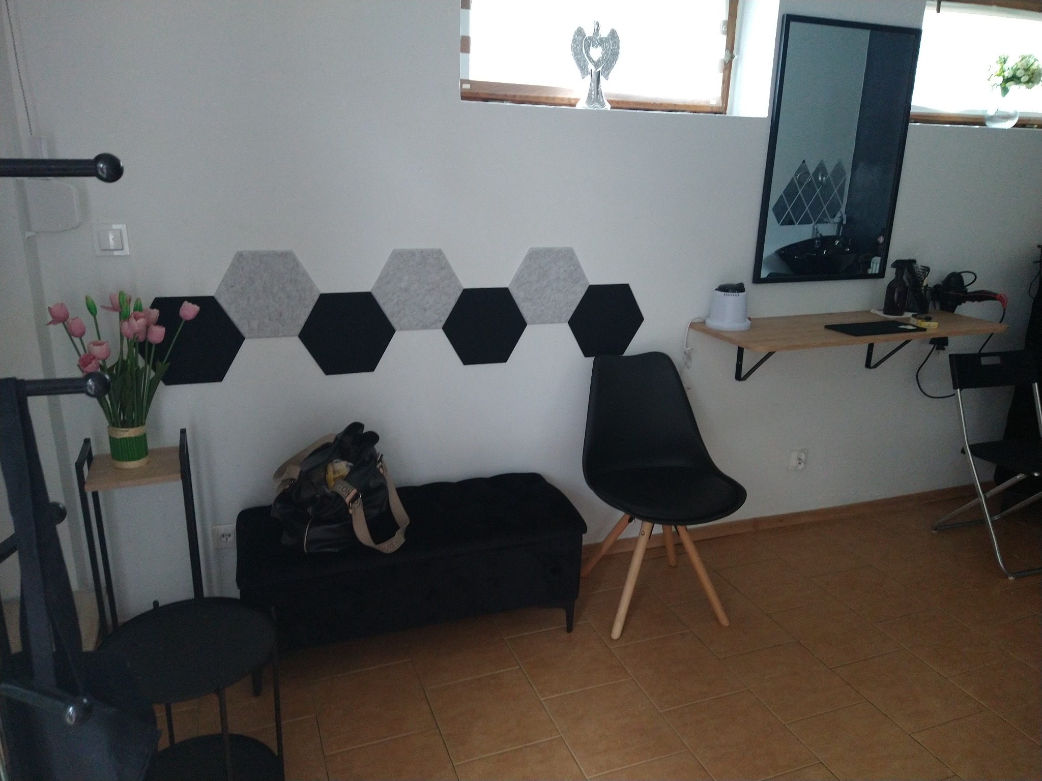 Wnętrze salonu z czarnym krzesłem, pufą i dekoracyjnymi panelami akustycznymi w kształcie sześciokątów na ścianie, lustro z odbiciem stanowiska i akcesoriów.