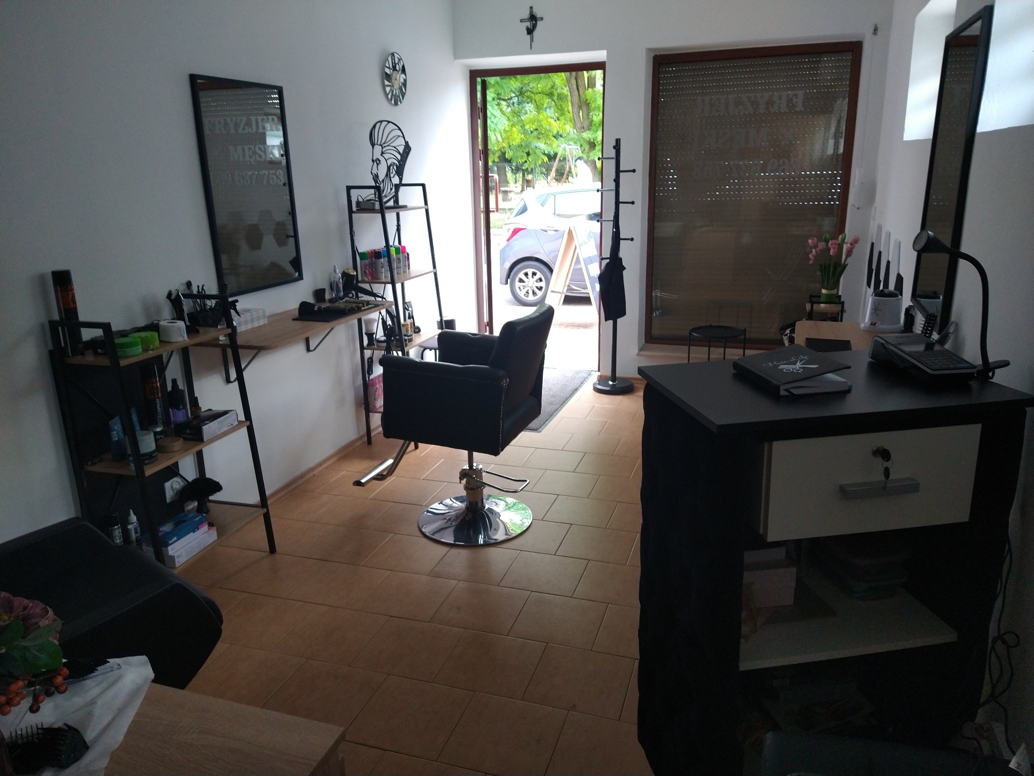 Wnętrze salonu fryzjerskiego z czarnym fotelem fryzjerskim na środku, widok na ulicę przez otwarte drzwi i lustro z napisem 'Fryzjer męski' na ścianie.
