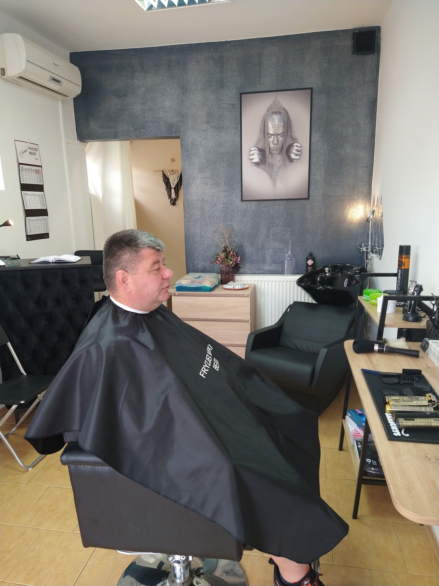 Mężczyzna w czarnej pelerynie siedzi na fotelu w salonie, oczekując na strzyżenie; w tle widoczna umywalka fryzjerska, narzędzia i plakat na ścianie.