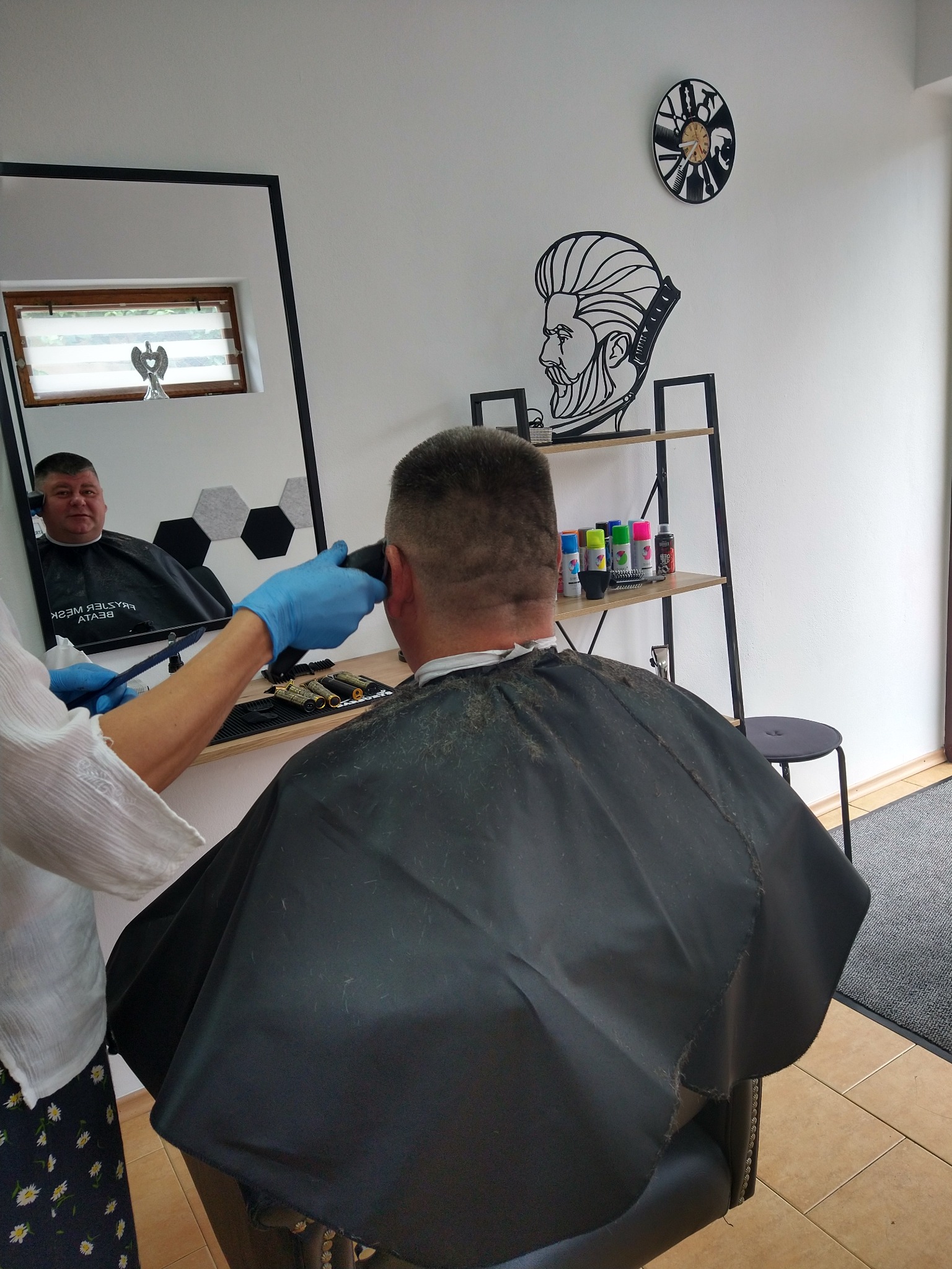 Strzyżenie męskie maszynką, widok z tyłu na klienta w czarnej pelerynie, detale wystroju salonu: zegar z motywem fryzjerskim i półka z kosmetykami do włosów.