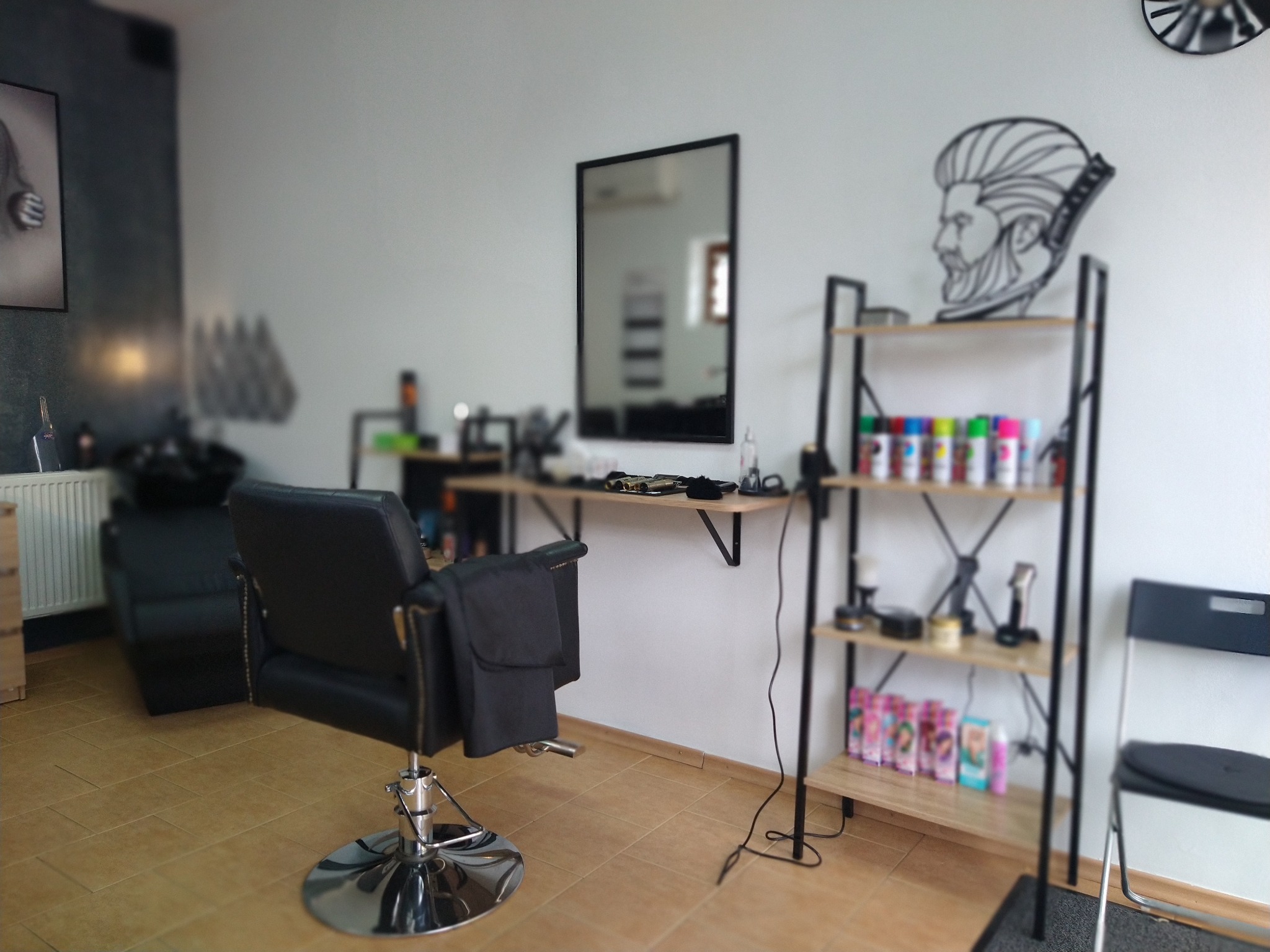 Wnętrze salonu z czarnym fotelem fryzjerskim na chromowanej podstawie, półką z kosmetykami i narzędziami, lustrem w czarnej ramie oraz dekoracją ścienną przedstawiającą profil brodatego mężczyzny.