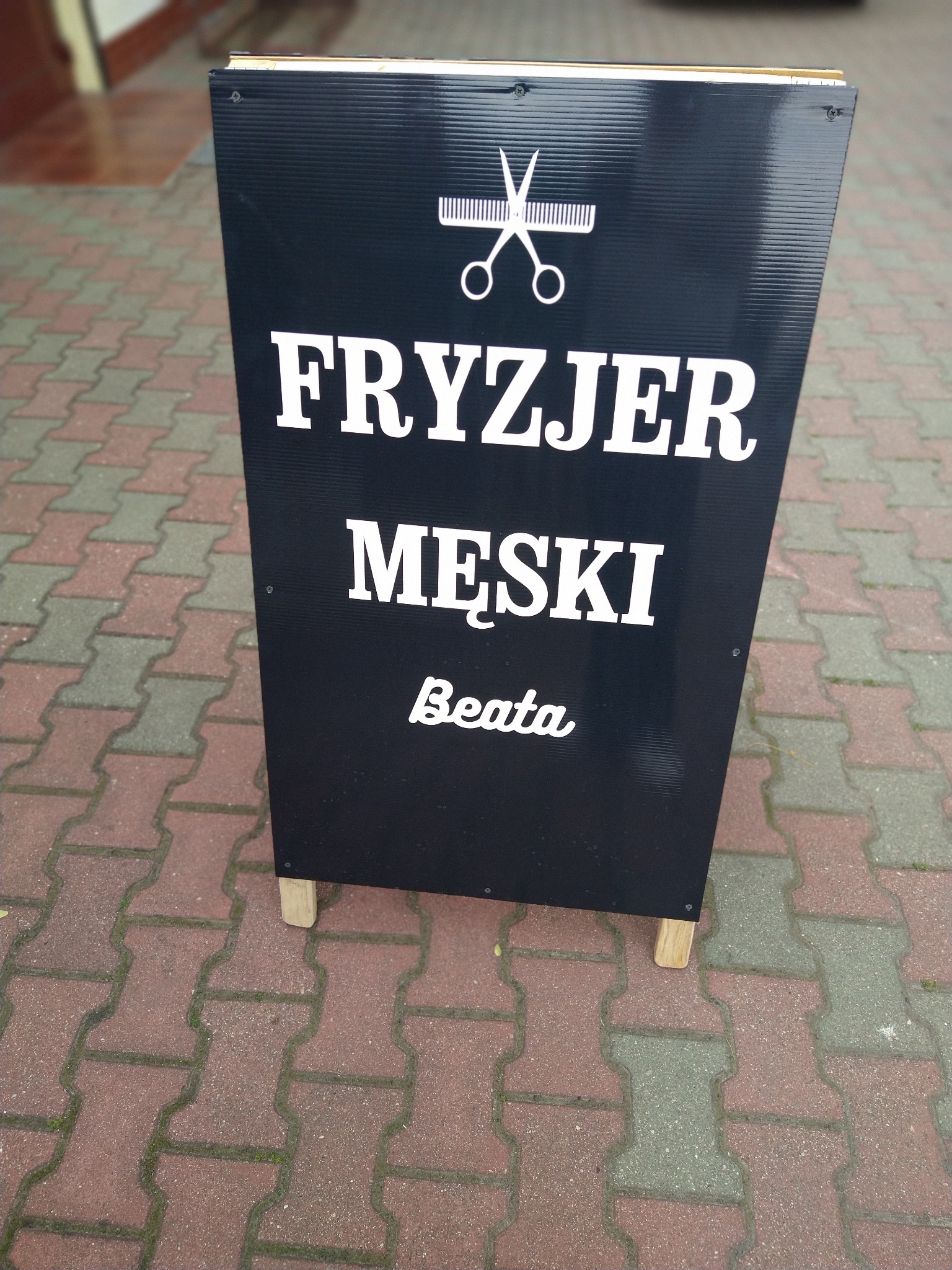 Czarny szyld reklamowy na drewnianych nóżkach, informujący o usługach strzyżenia męskiego, z grafiką nożyczek i grzebienia, ustawiony na chodniku z kostki.