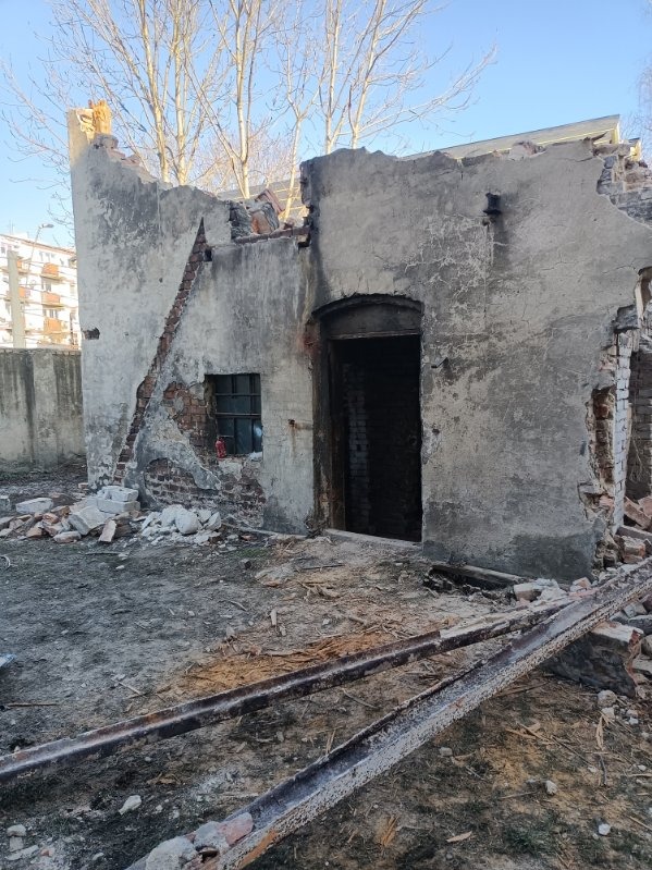 Częściowo rozebrany budynek z widocznymi fragmentami cegieł i resztkami ścian, z metalowymi elementami leżącymi na ziemi, okno z kratą, w tle blok mieszkalny.