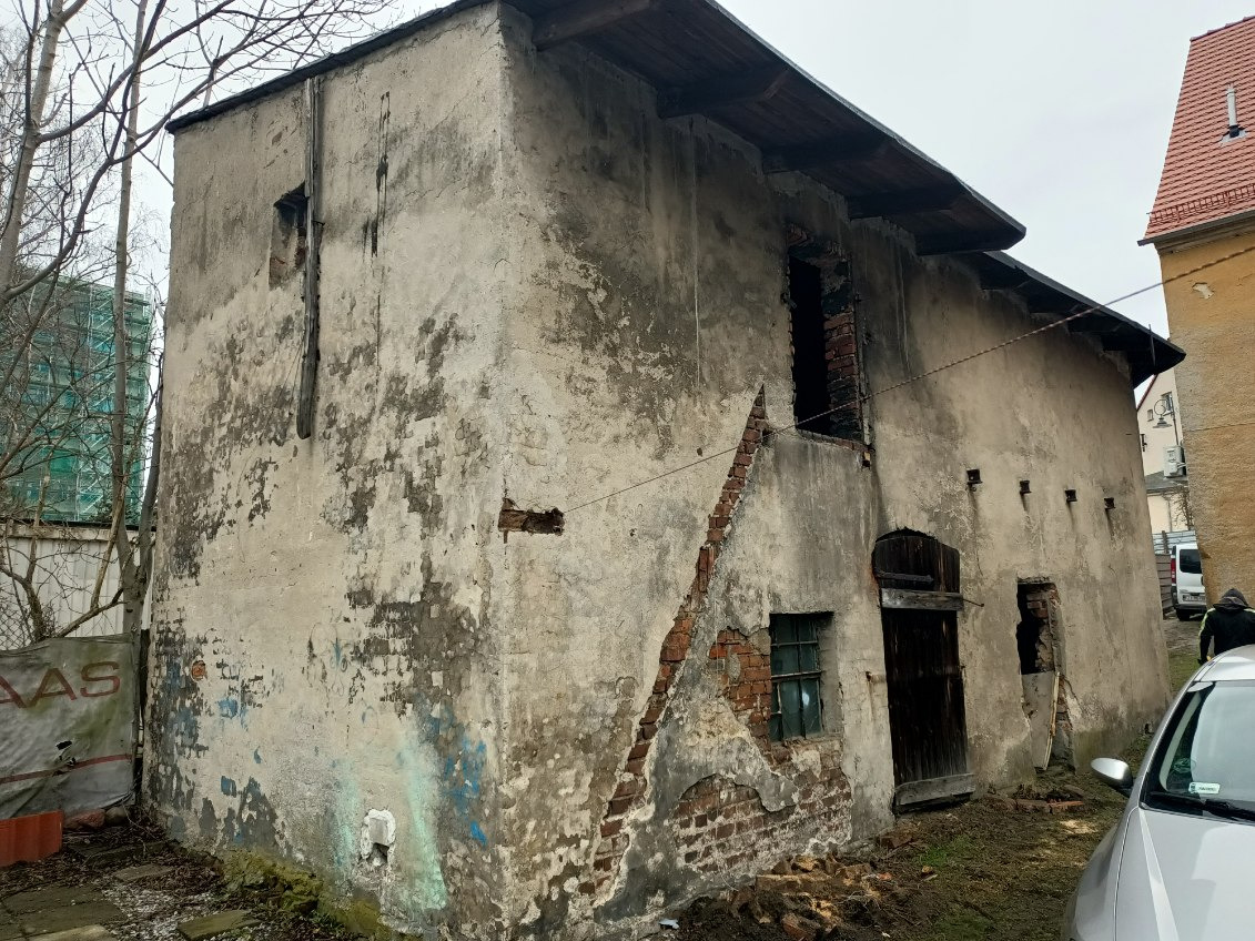 Stary, zaniedbany budynek z uszkodzoną elewacją, widoczne fragmenty cegieł, wybite okna i drzwi, zarośnięty teren wokół, częściowo zaparkowany samochód z przodu.