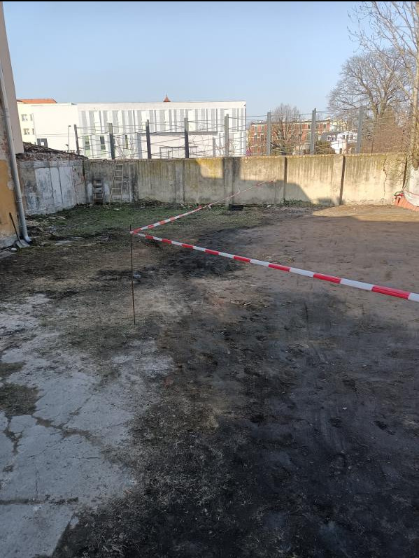 Teren po wyburzeniu z fragmentami ścian i ogrodzeniem, oznaczony taśmą ostrzegawczą w kolorze biało-czerwonym, widoczna drabina oparta o mur i budynek w tle.