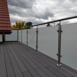 Volodymyr Mikhalets Amix balustrady nierdzewne - Matowa szklana balustrada z nierdzewnymi słupkami na tarasie z desek kompozytowych, widok na pochmurne niebo i fragment dachu sąsiedniego budynku.