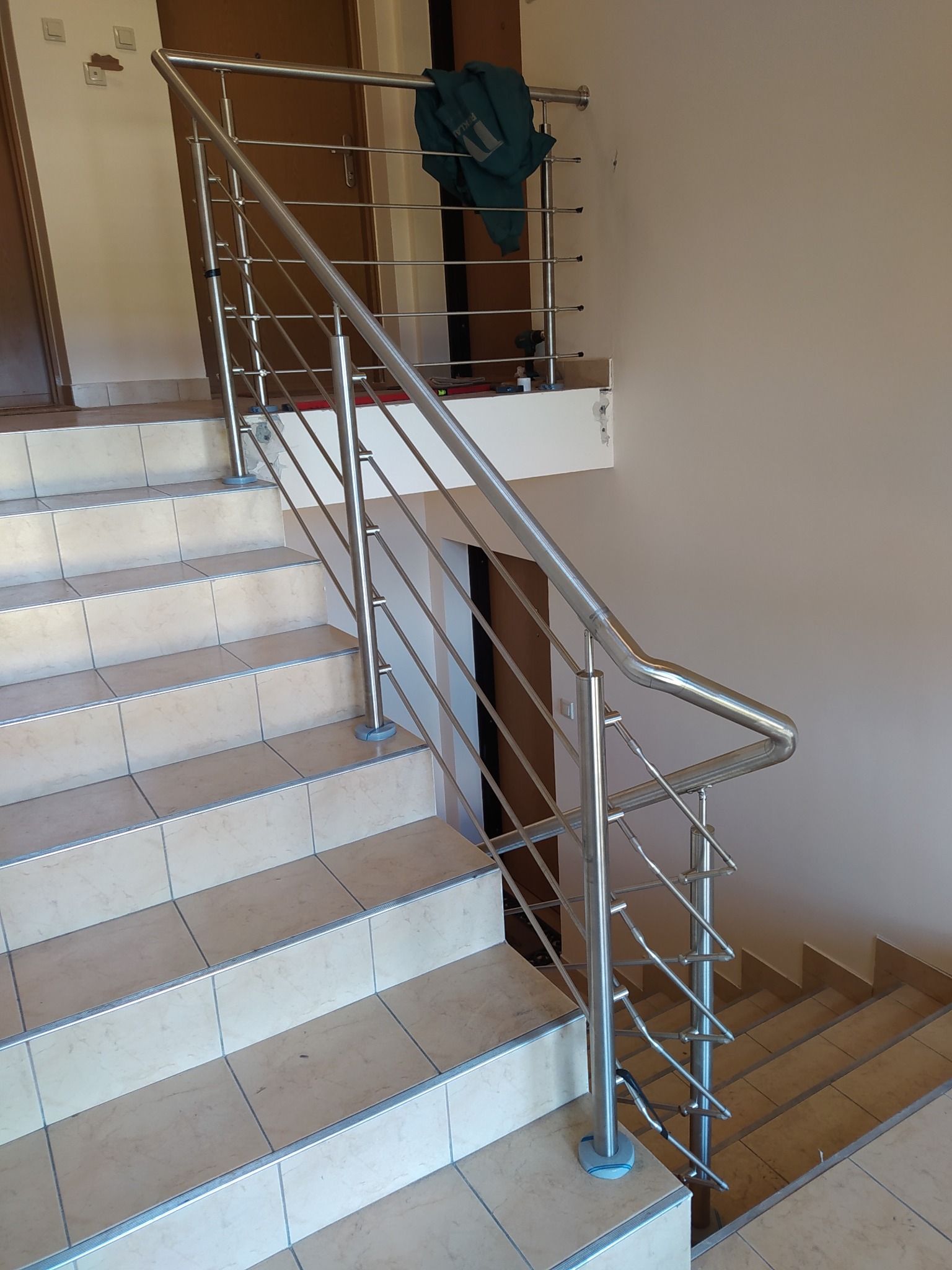 Polerowana, metalowa balustrada schodowa z pionowymi prętami, zamocowana do beżowych, kwadratowych płytek schodowych. Na półpiętrze widoczna zielona bluza i narzędzia.
