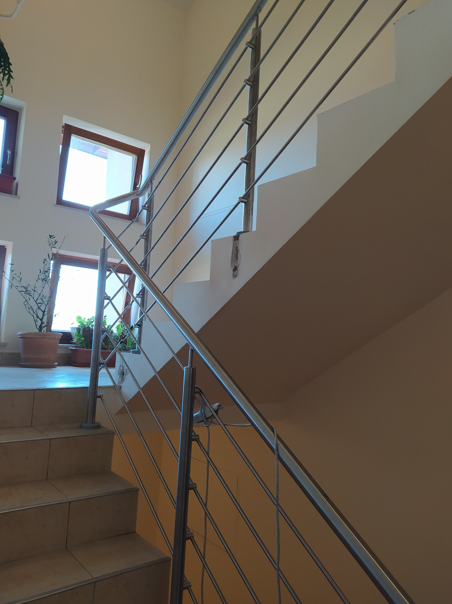 Nowoczesna klatka schodowa z jasnymi płytkami i minimalistyczną, metalową poręczą z poziomymi prętami, widok z dołu na tle okien z roślinami.