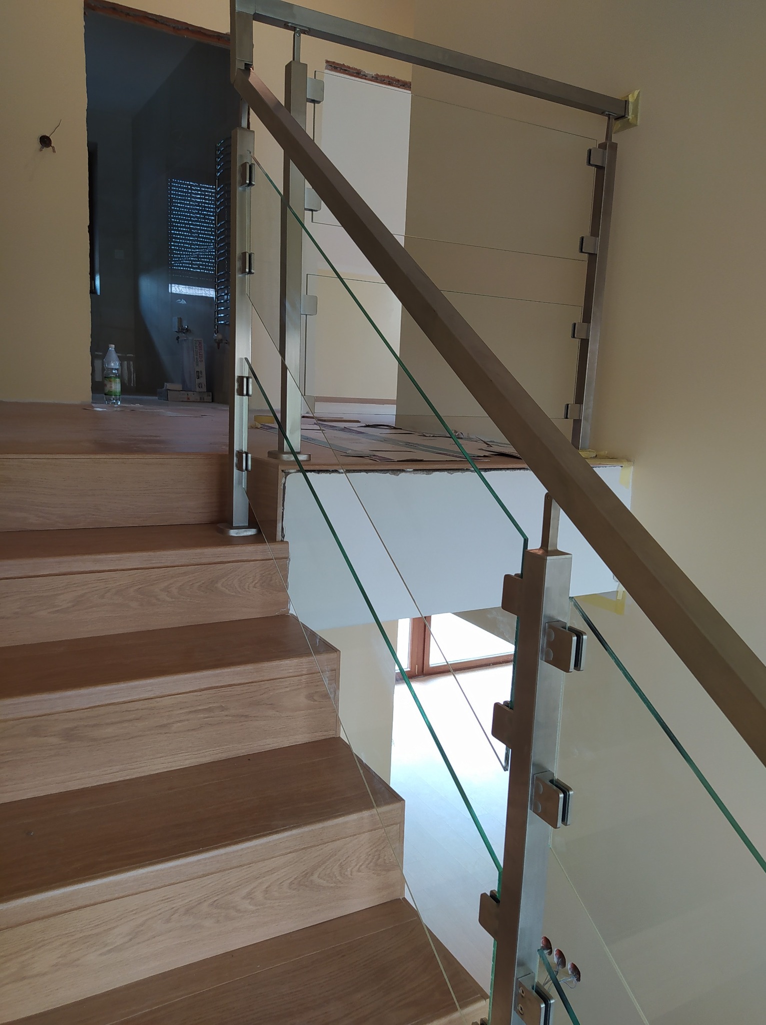 Szczegółowe ujęcie schodów z drewnianymi stopniami i balustradą szklaną mocowaną do słupków ze stali nierdzewnej; widoczne niedokończone prace wykończeniowe w tle.