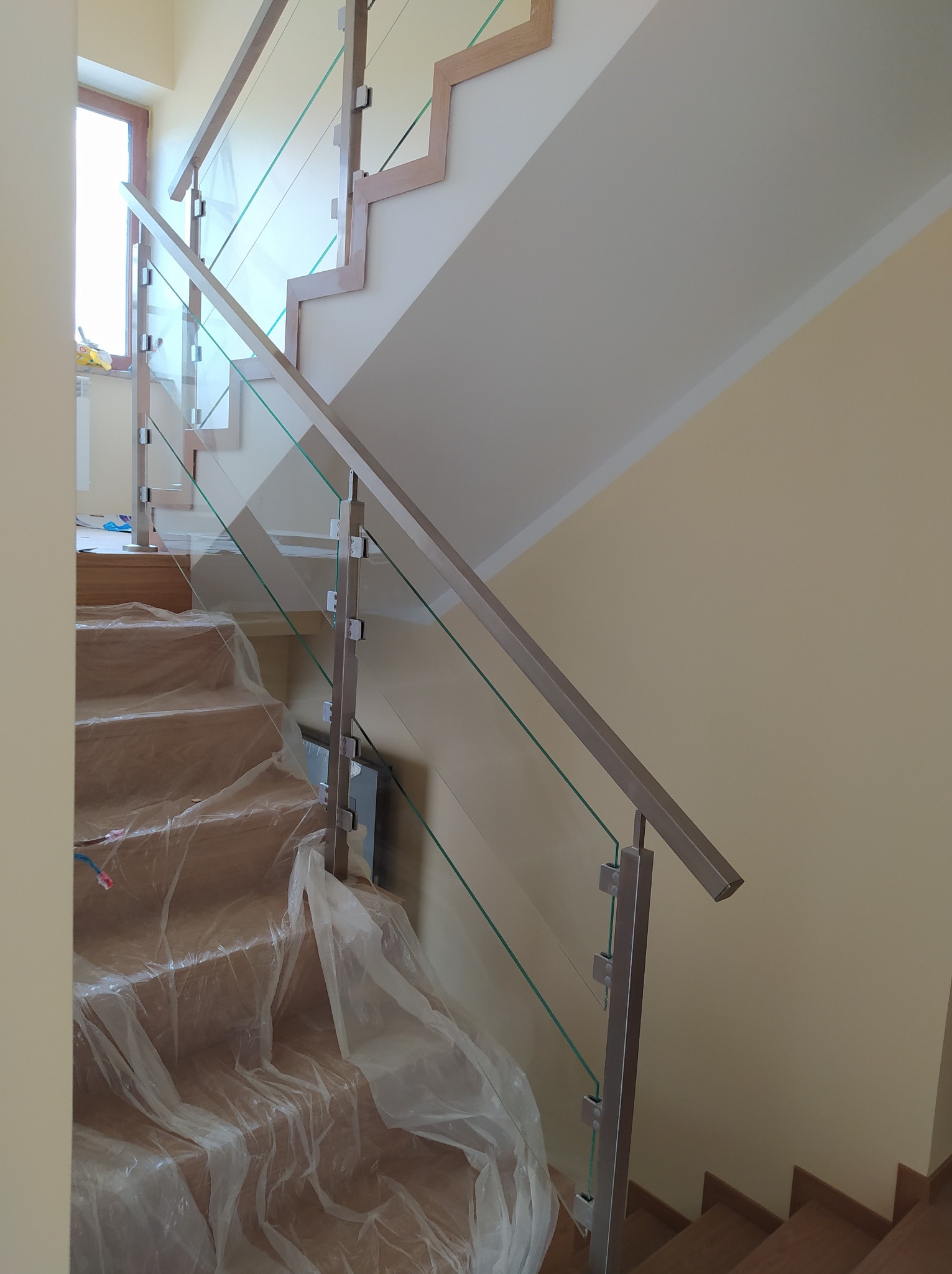 Szklana balustrada ze stalowymi słupkami i poręczą na drewnianych schodach, zabezpieczonych folią ochronną podczas montażu.