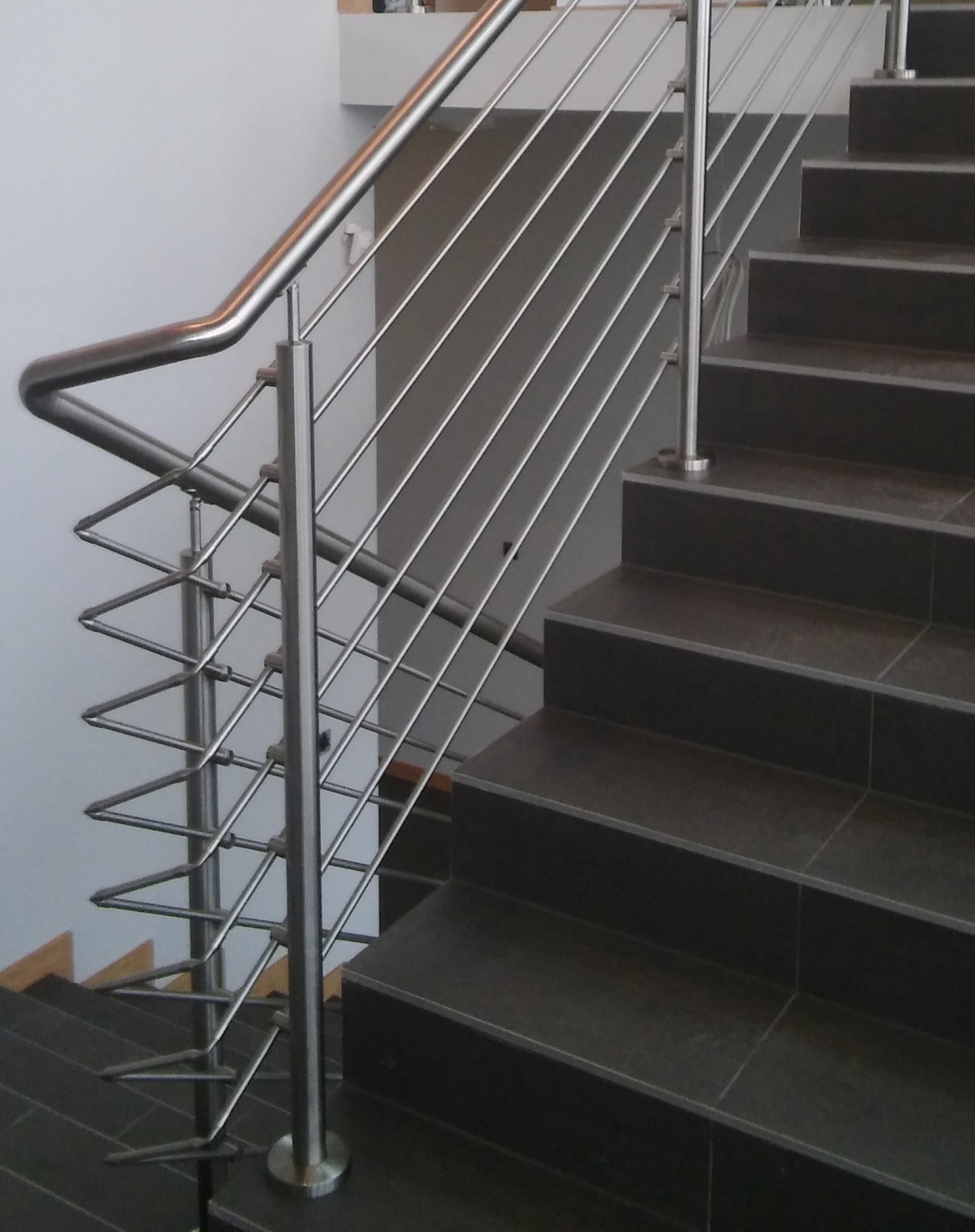 Schody wyłożone ciemnymi płytkami z balustradą ze stali nierdzewnej z wypełnieniem z cienkich prętów, mocowanych w nietypowy zygzakowaty sposób.