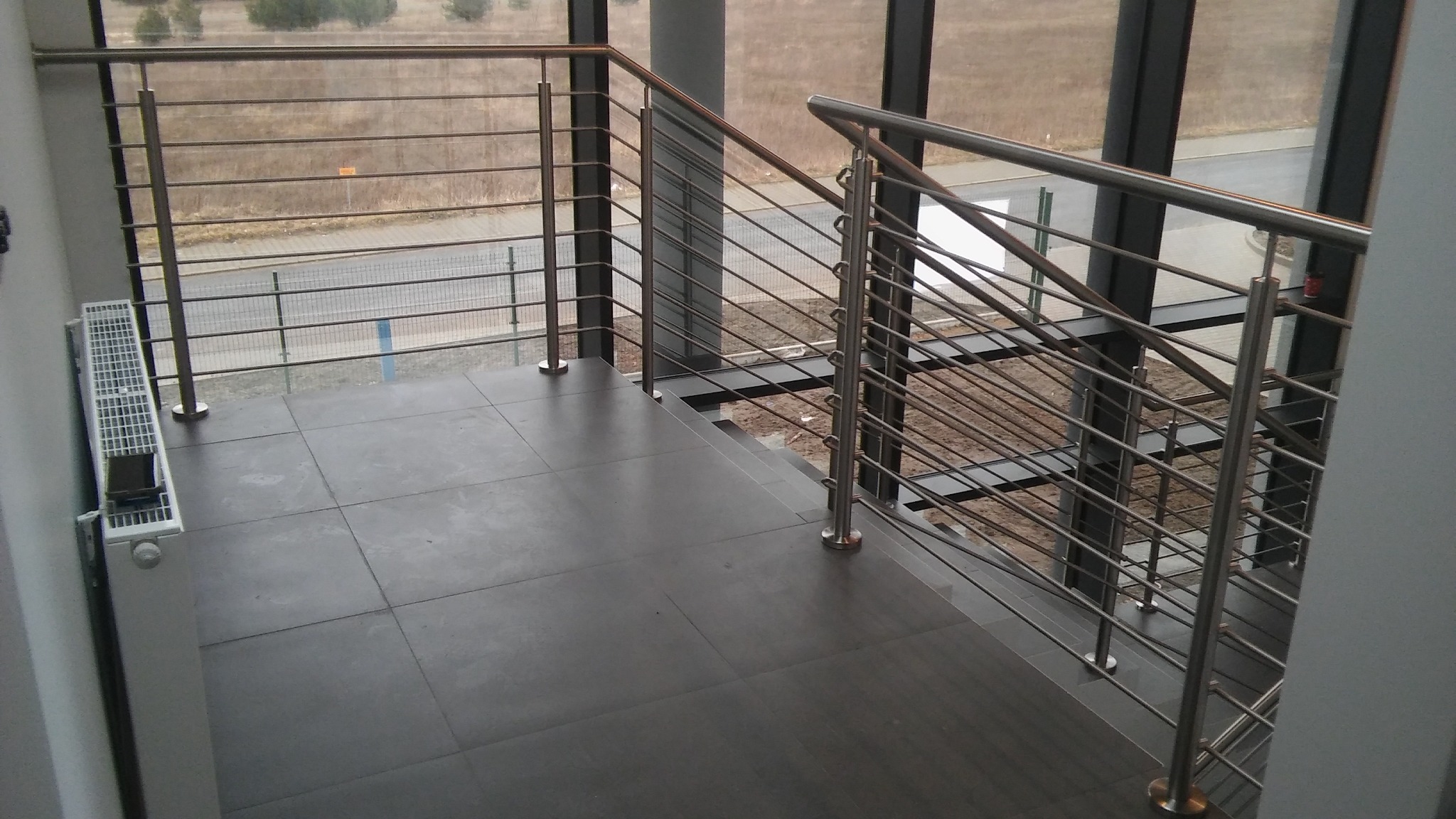 Schody z szarymi płytkami i metalową balustradą z poziomymi prętami, widok z góry na zewnątrz przez duże okno.