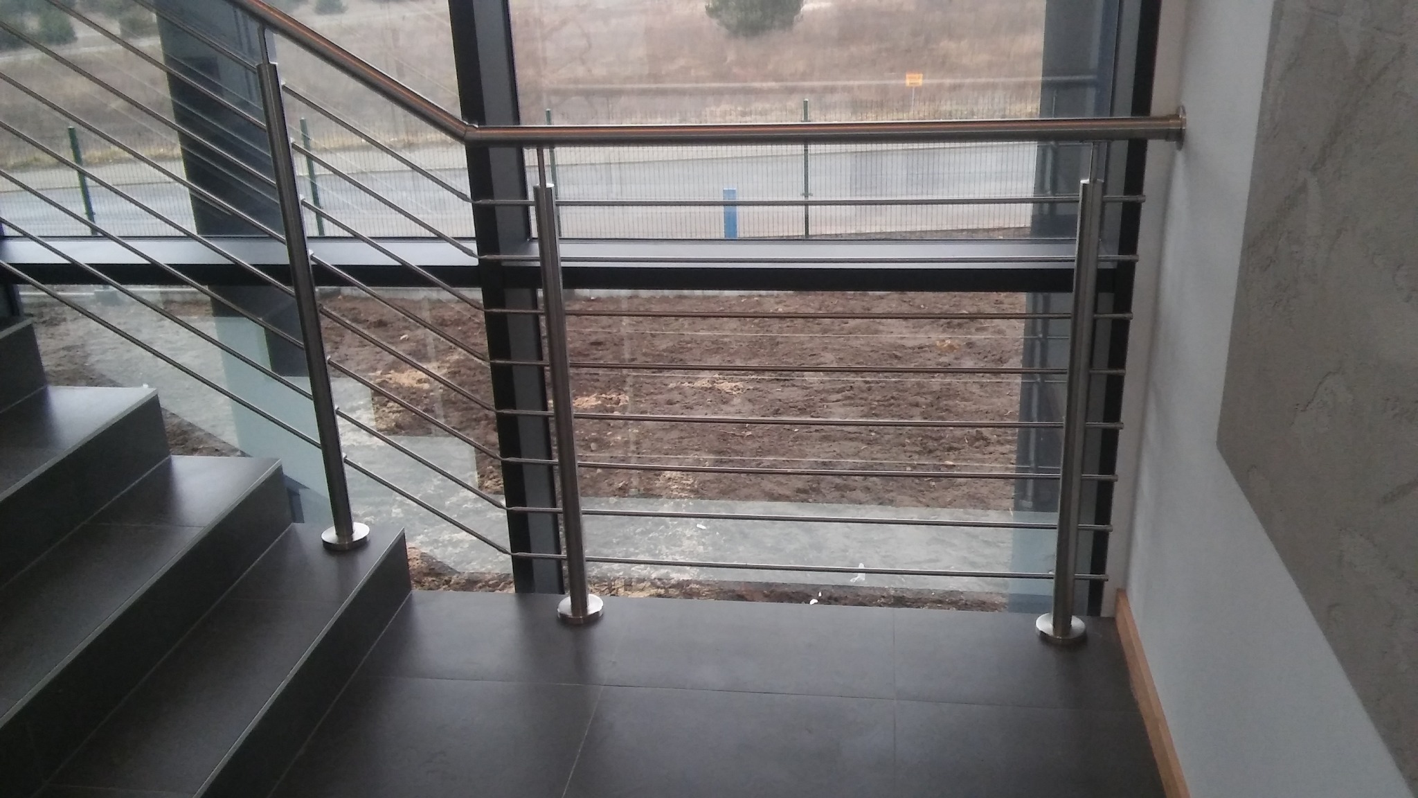 Wewnętrzna klatka schodowa z ciemnymi schodami i nowoczesną balustradą ze stali nierdzewnej z poziomymi prętami, widok przez okno na zewnątrz.