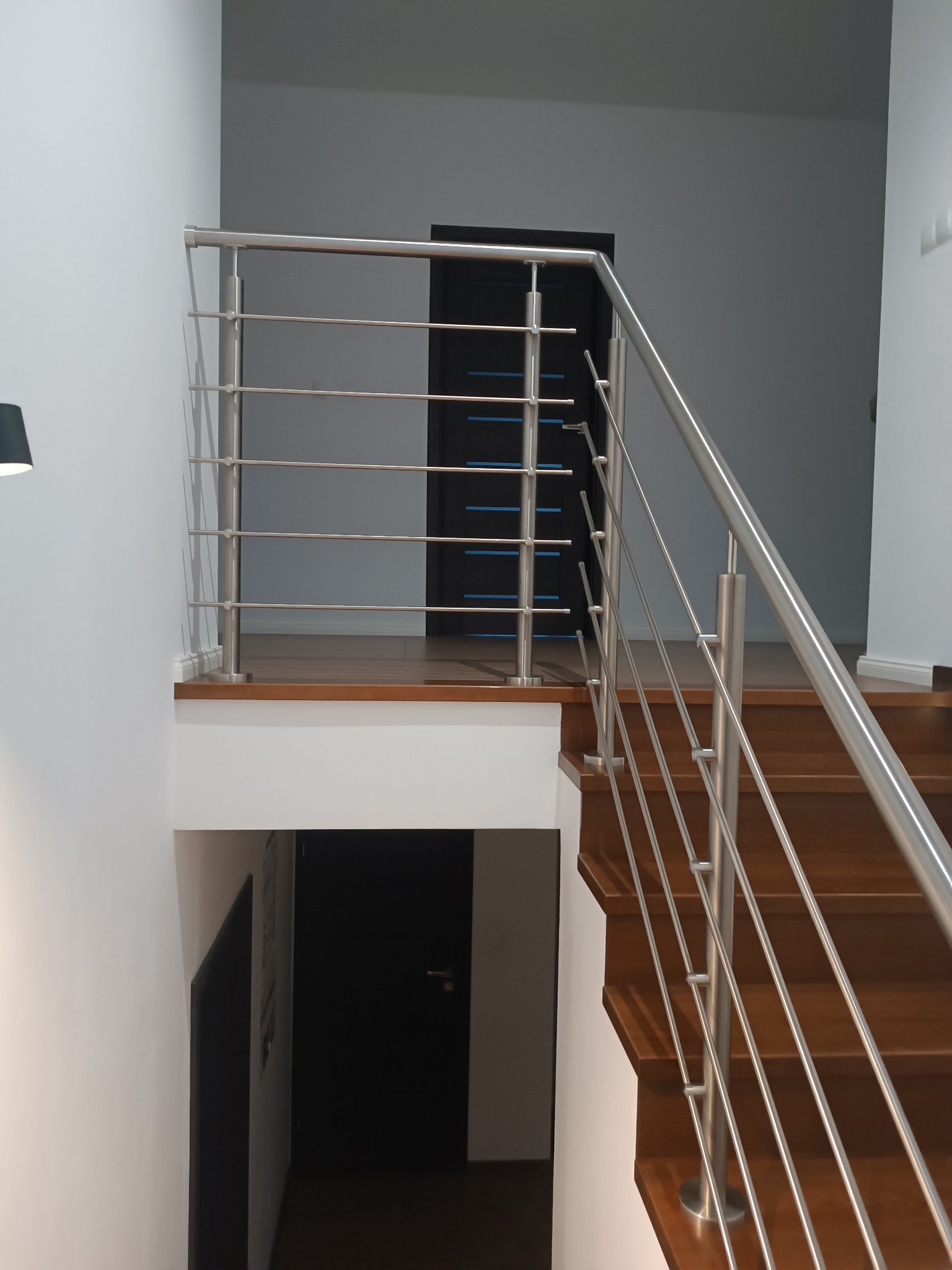 Minimalistyczna balustrada ze stali nierdzewnej z poziomymi prętami, zamontowana na drewnianych schodach w nowoczesnym wnętrzu z białymi ścianami i ciemnymi drzwiami.