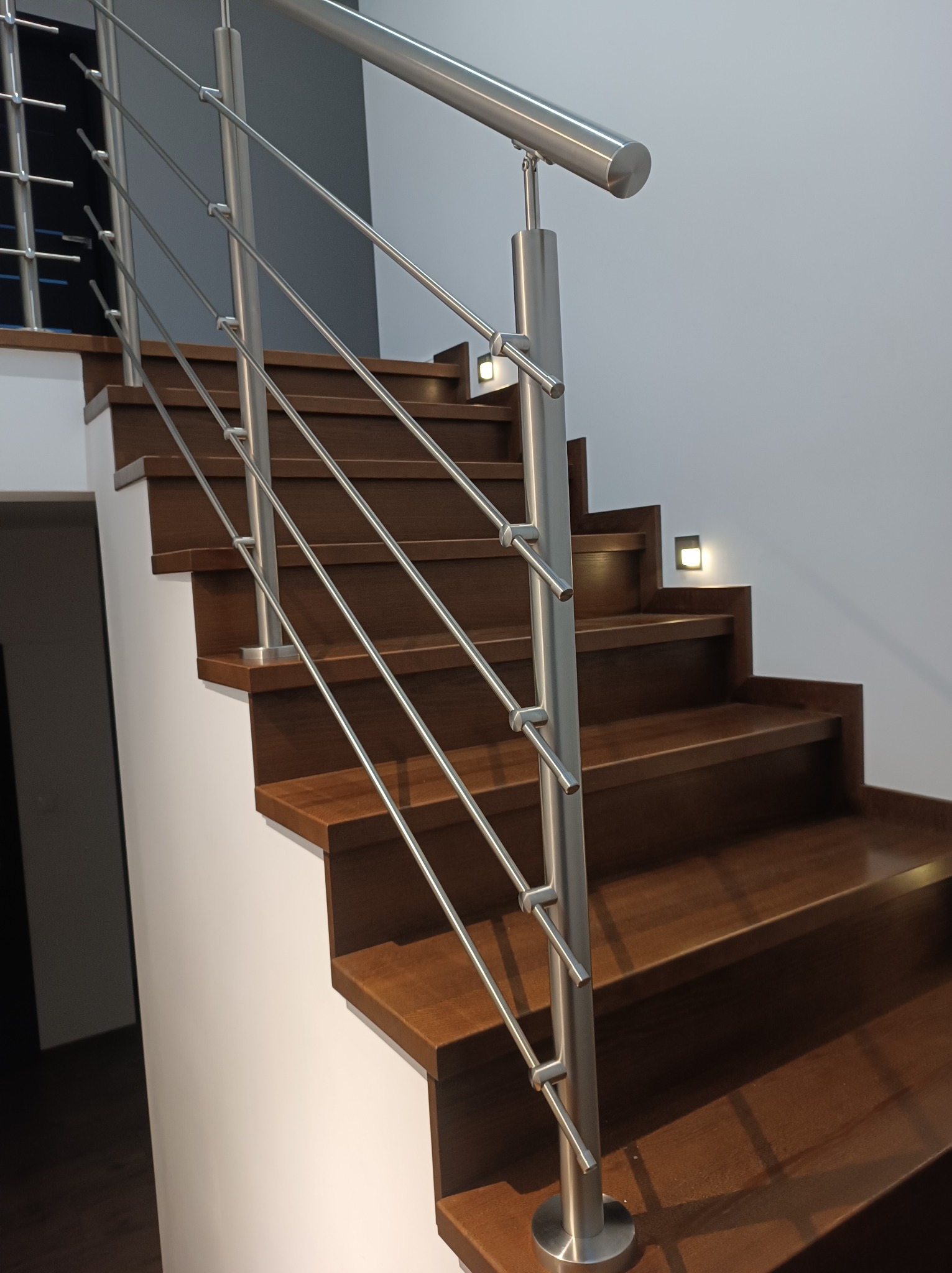 Nowoczesne schody z ciemnego drewna z minimalistyczną balustradą ze stali nierdzewnej, oświetlenie punktowe na ścianie.