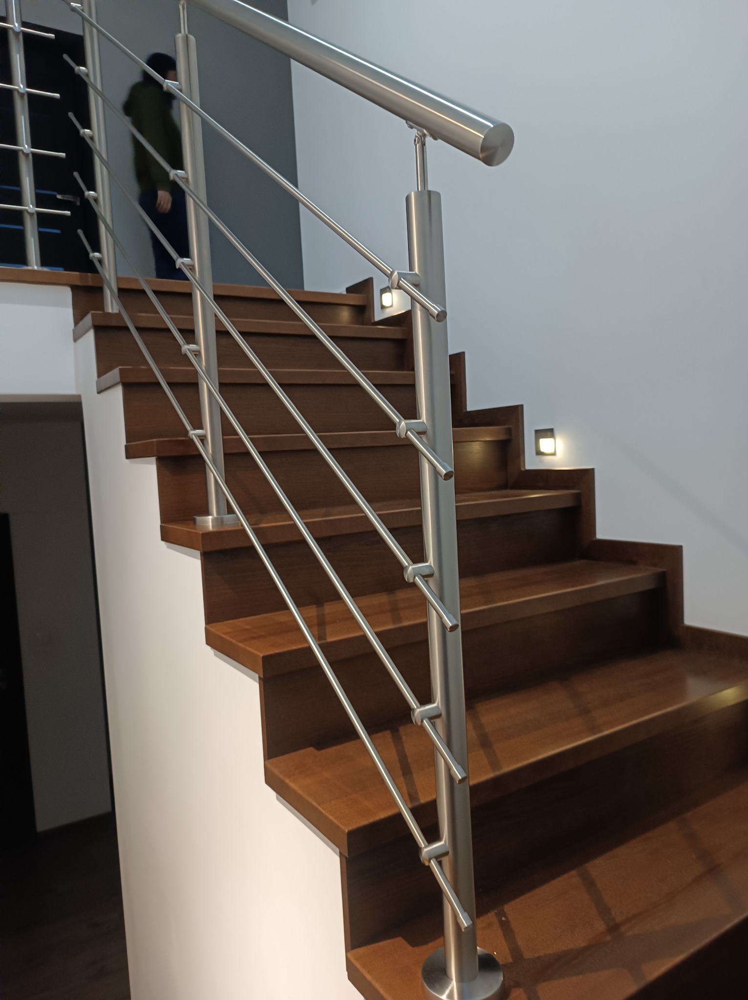 Schody z ciemnego drewna z nowoczesną balustradą ze stali nierdzewnej, oświetlenie schodowe, widoczna osoba w tle.