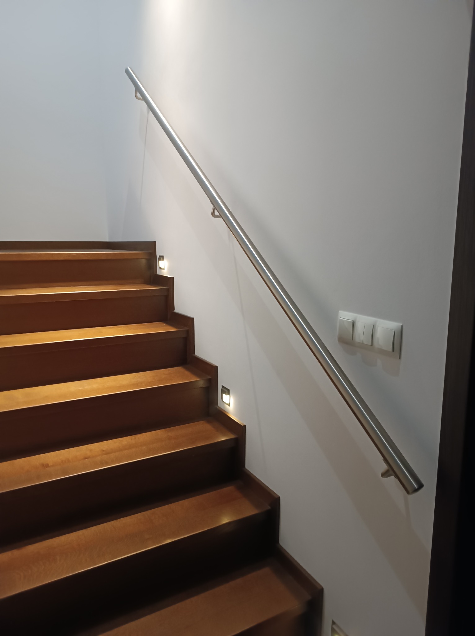 Schody drewniane z oświetleniem LED w stopniach i stalowa poręcz zamontowana do ściany, widoczny włącznik światła.