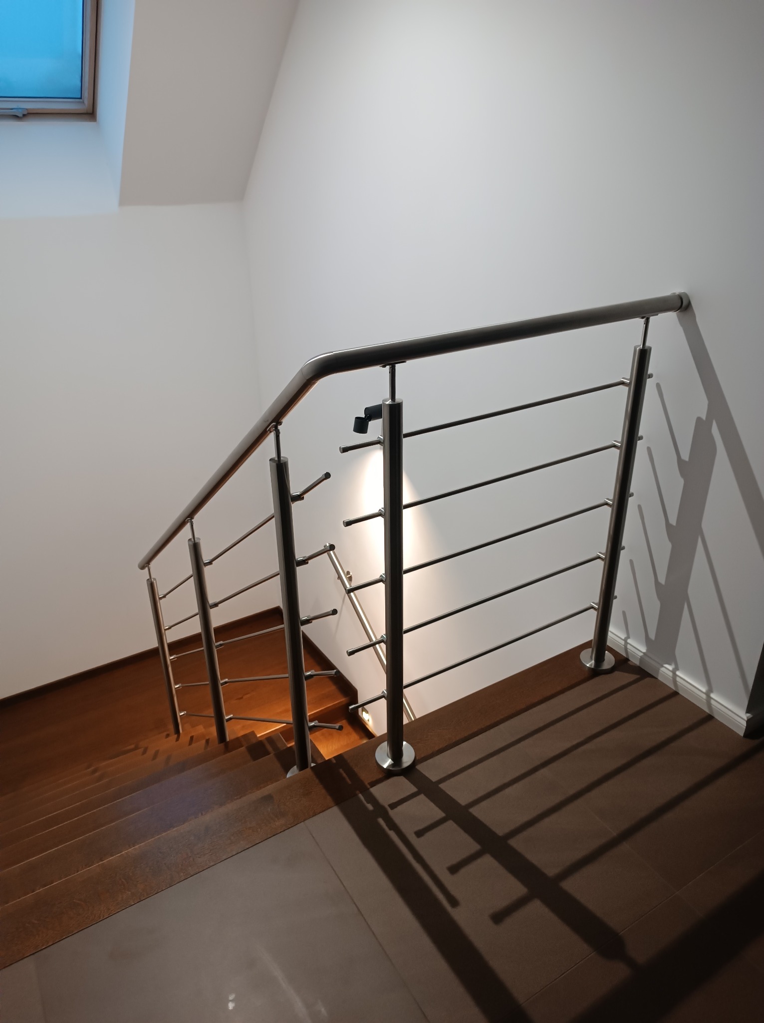 Schody z brązowymi stopniami i balustradą nierdzewną z poziomymi poprzeczkami, widok z góry, oświetlenie od okna dachowego.