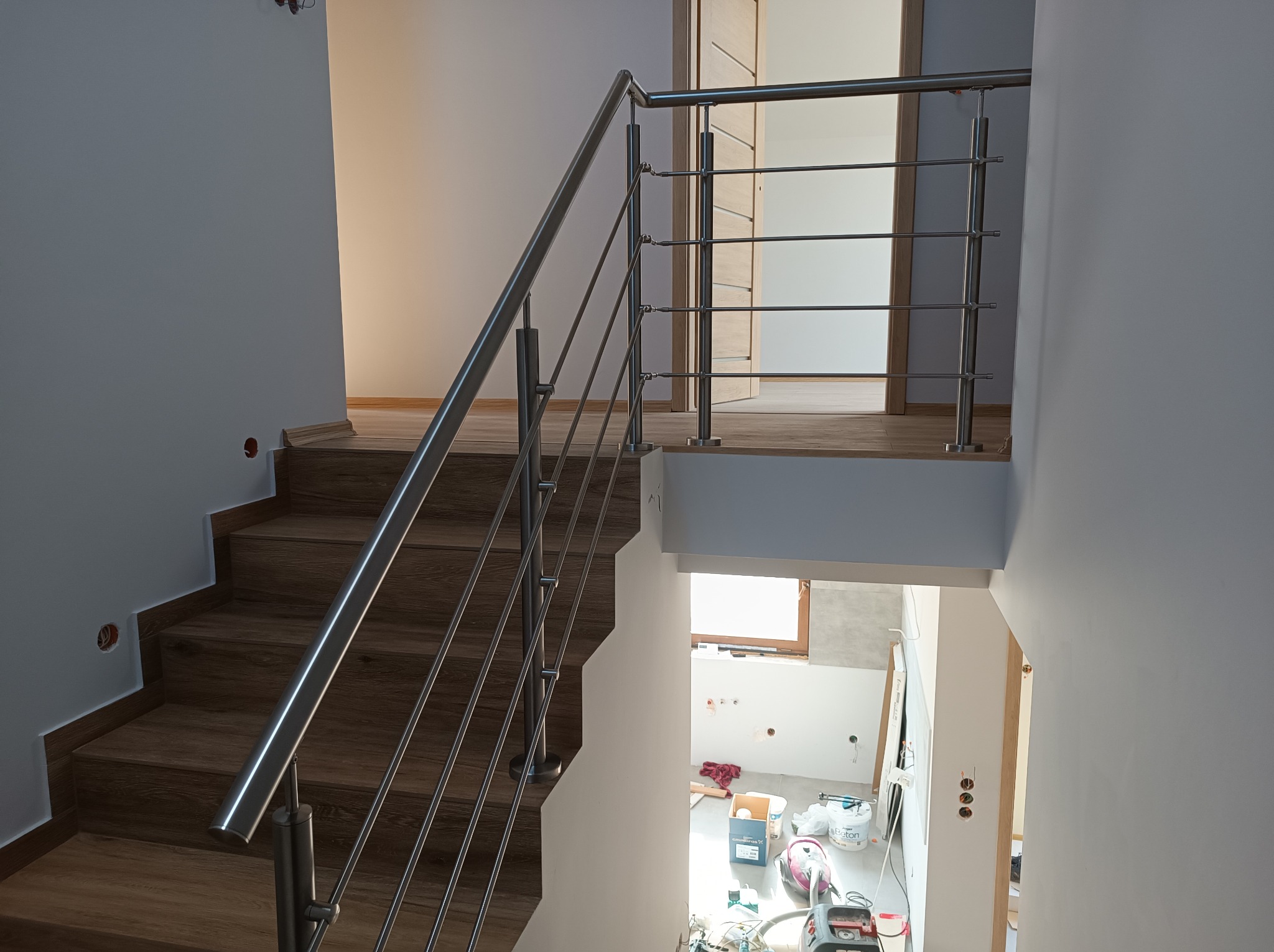 Wnętrze domu w trakcie wykańczania: drewniane schody z nowoczesną, poziomą, metalową poręczą, widok z góry na dół, z widocznym pomieszczeniem w trakcie remontu.