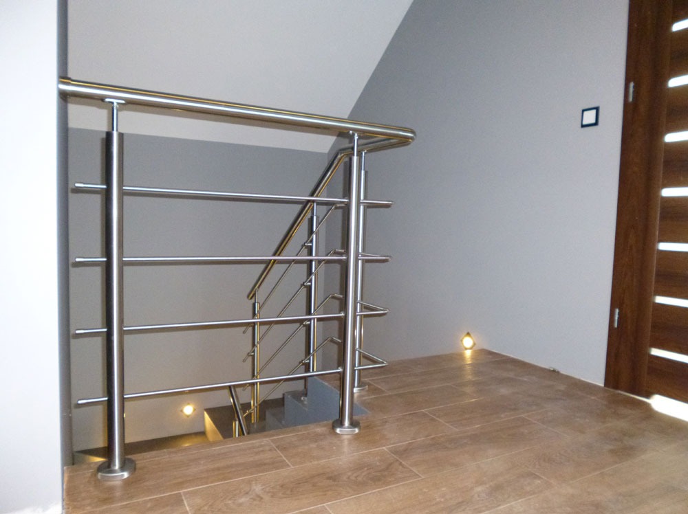 Polerowana, metalowa balustrada z poziomymi prętami wypełniającymi, zamontowana przy schodach w nowoczesnym wnętrzu z drewnopodobną podłogą i punktowym oświetleniem przypodłogowym.