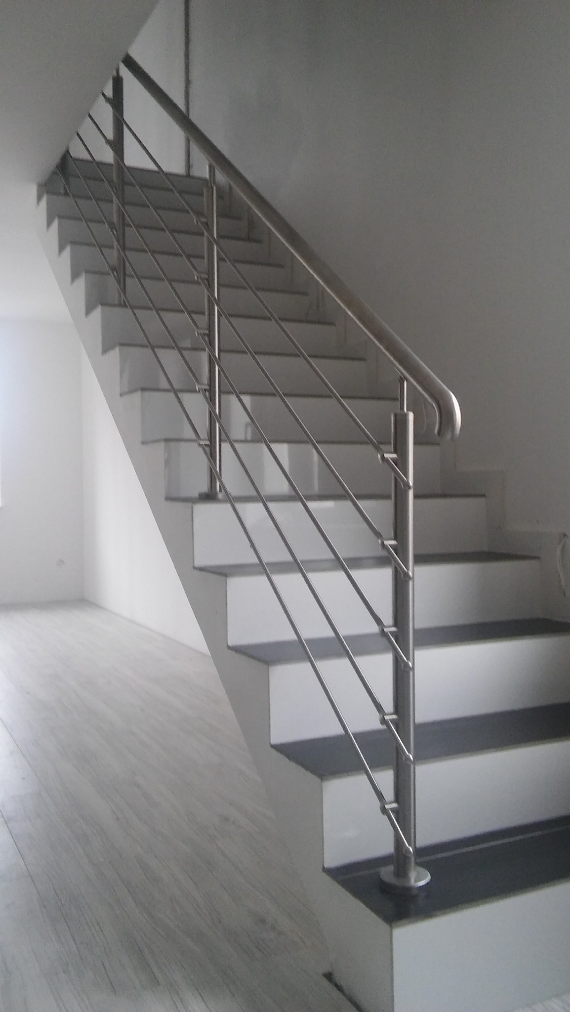 Nierdzewna balustrada schodowa z wypełnieniem prętowym, widok z dołu schodów, minimalistyczne wnętrze w odcieniach szarości.