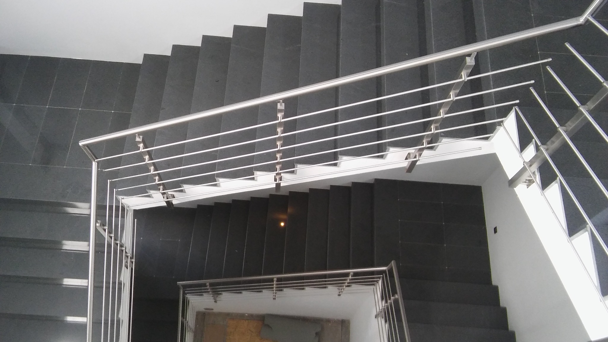 Widok z góry na schody z czarnego kamienia z prostą, nowoczesną metalową balustradą z poziomymi prętami, zabezpieczającą przestrzeń między piętrami.