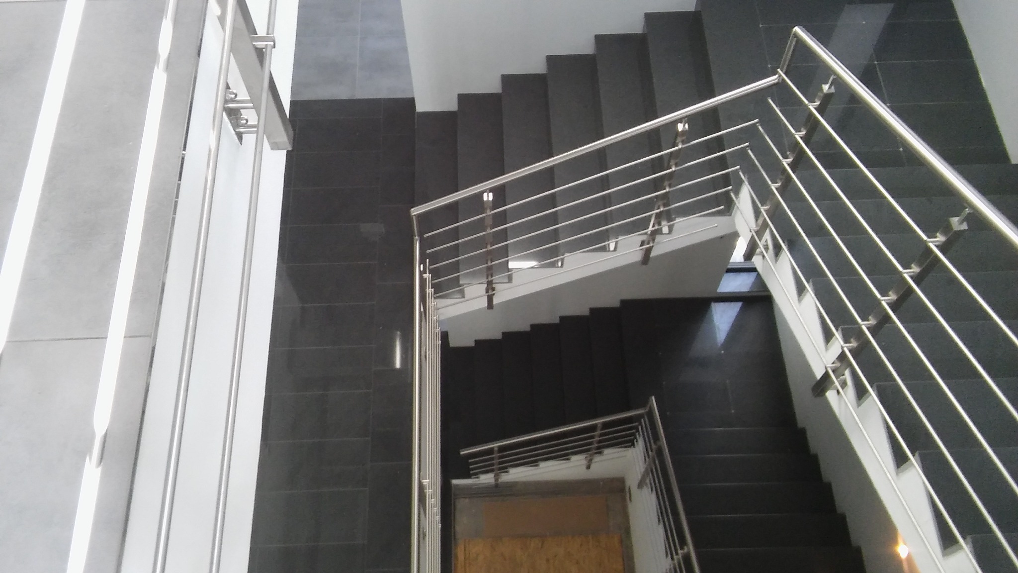 Widok z góry na klatkę schodową z czarnymi schodami i metalowymi poręczami z poziomymi szczeblami, geometryczny układ linii i kątów.