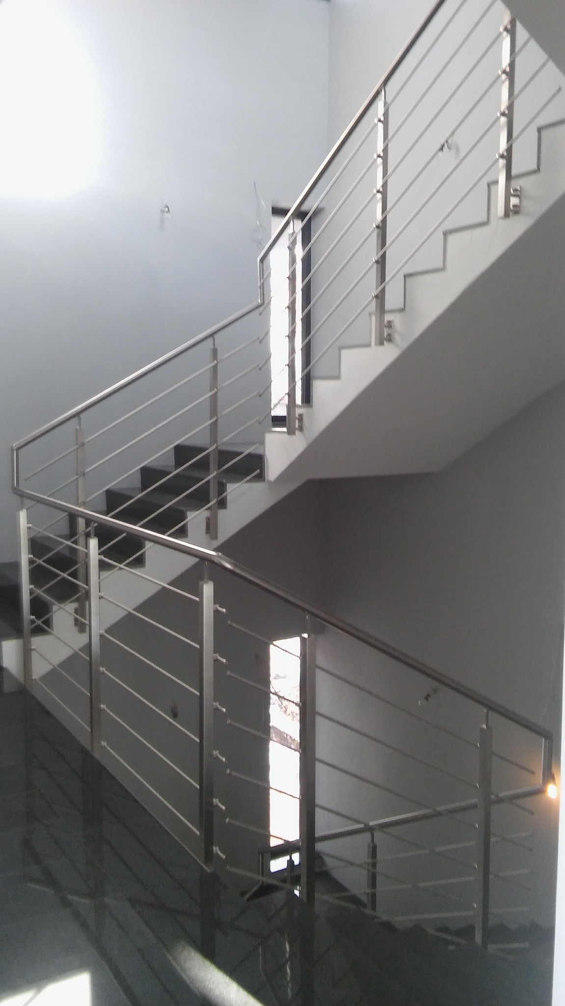 Schody wewnętrzne z błyszczącą posadzką i minimalistyczną balustradą ze stali nierdzewnej z poziomymi prętami, oświetlone wpadającym światłem z okna.