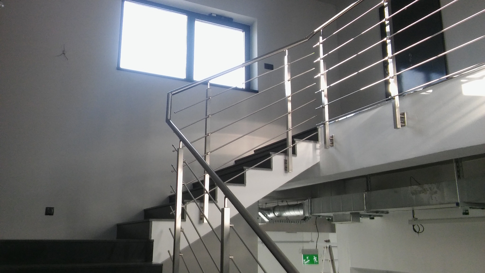 Nowoczesne schody z metalowymi poręczami i minimalistyczną balustradą ze stali nierdzewnej, widoczne elementy wentylacji pod schodami, jasne wnętrze z oknem na górze.