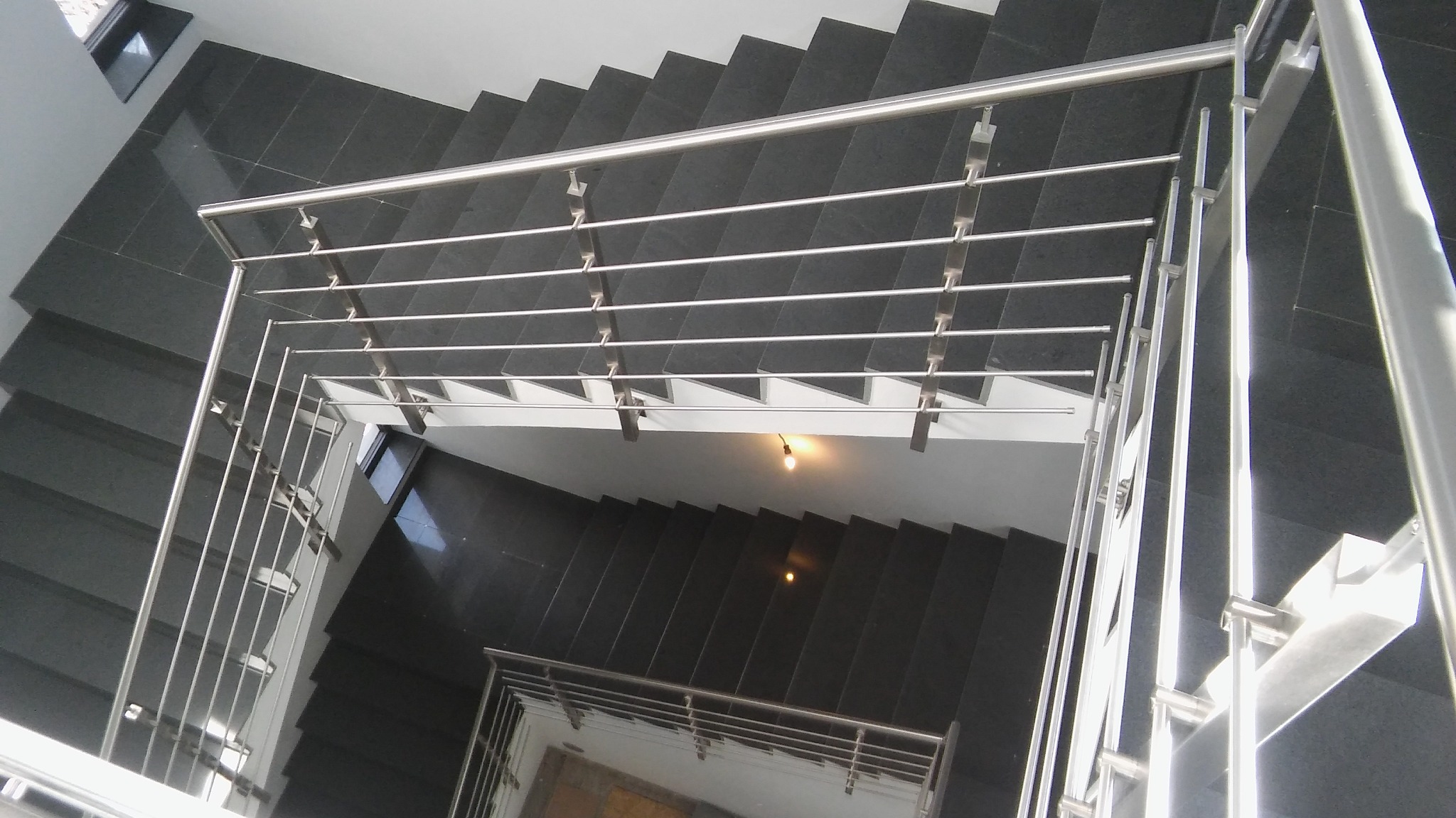 Widok z góry na schody z czarnego kamienia z nowoczesną balustradą ze stali nierdzewnej z poziomymi prętami, oświetlone żarówką.