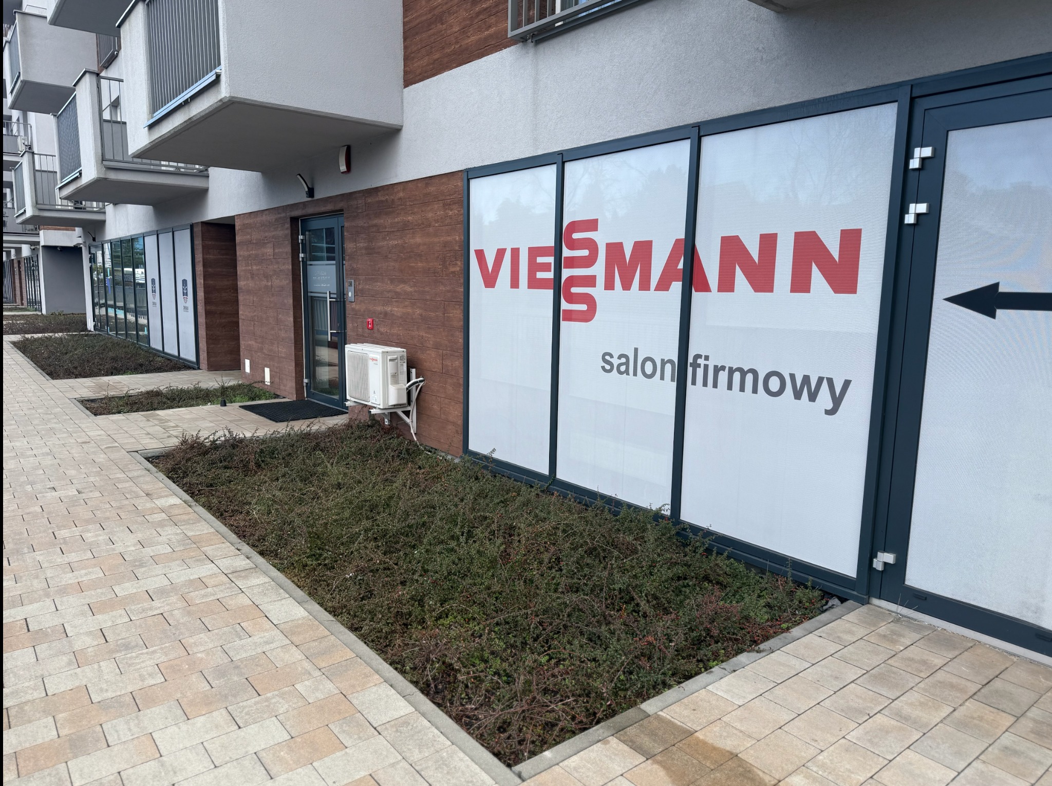 Fasada budynku z dużym oknem wystawowym prezentującym logo i napis 'salon firmowy' marki Viessmann, z klimatyzatorem na ścianie obok wejścia.