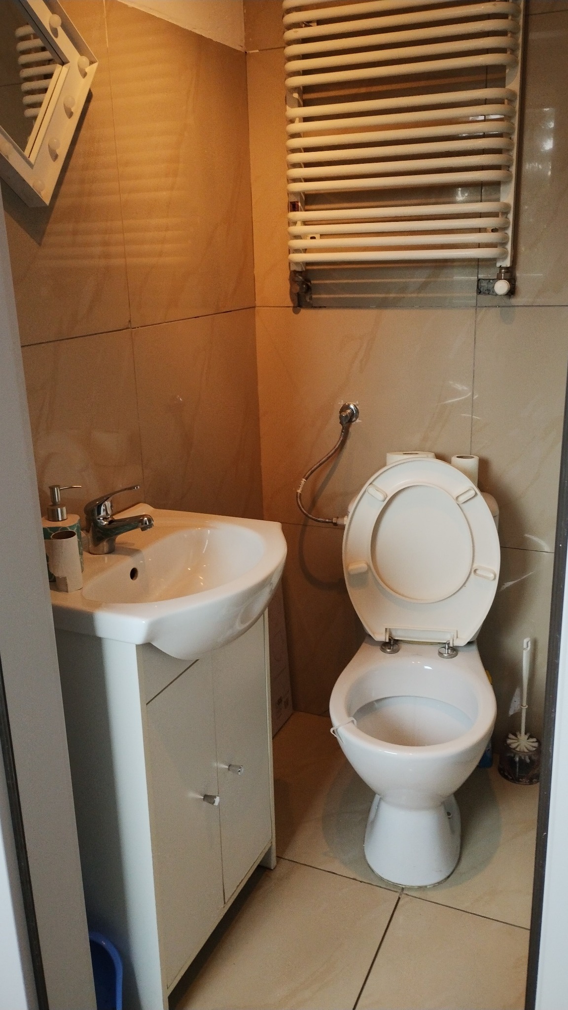 Umywalka z szafką, toaleta z otwartą klapą i grzejnik drabinkowy w małej łazience wyłożonej beżowymi płytkami.