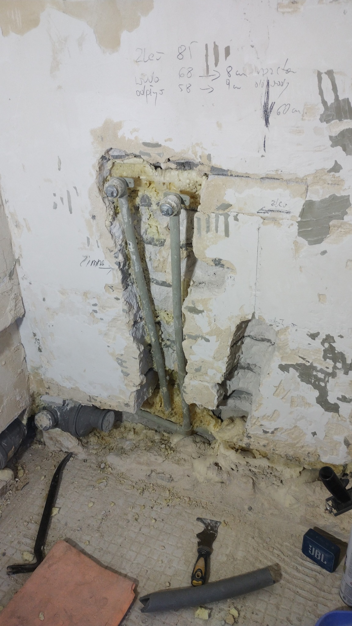 Odkryte rury wodne w trakcie remontu łazienki, widoczne oznaczenia na ścianie, gruz i narzędzia na podłodze.