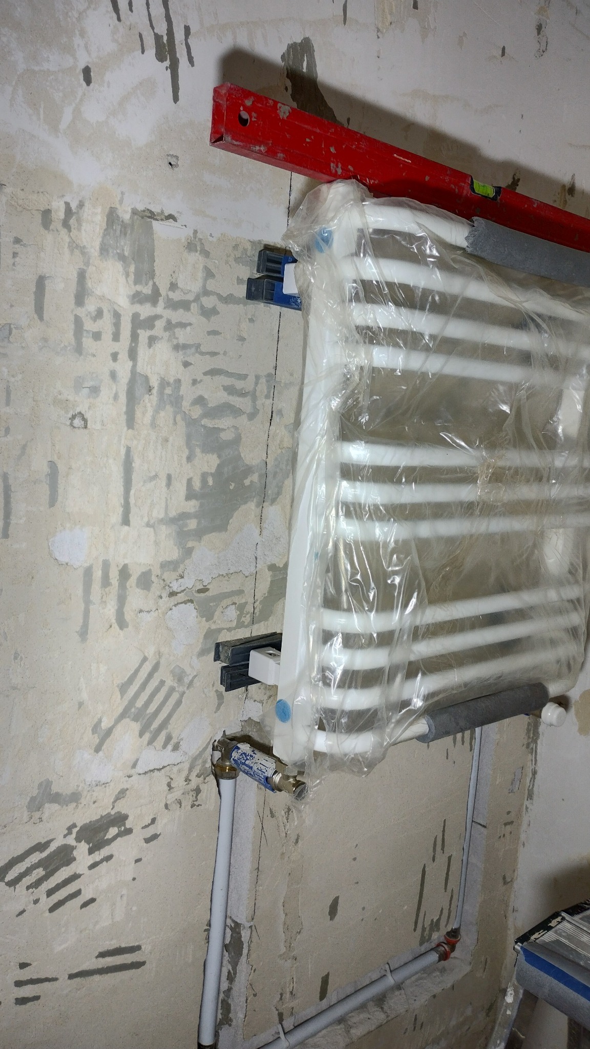 Grzejnik drabinkowy, zabezpieczony folią, zamontowany na ścianie w trakcie remontu łazienki, widoczne rury, zawory, poziomica i ślady po wcześniejszych płytkach.
